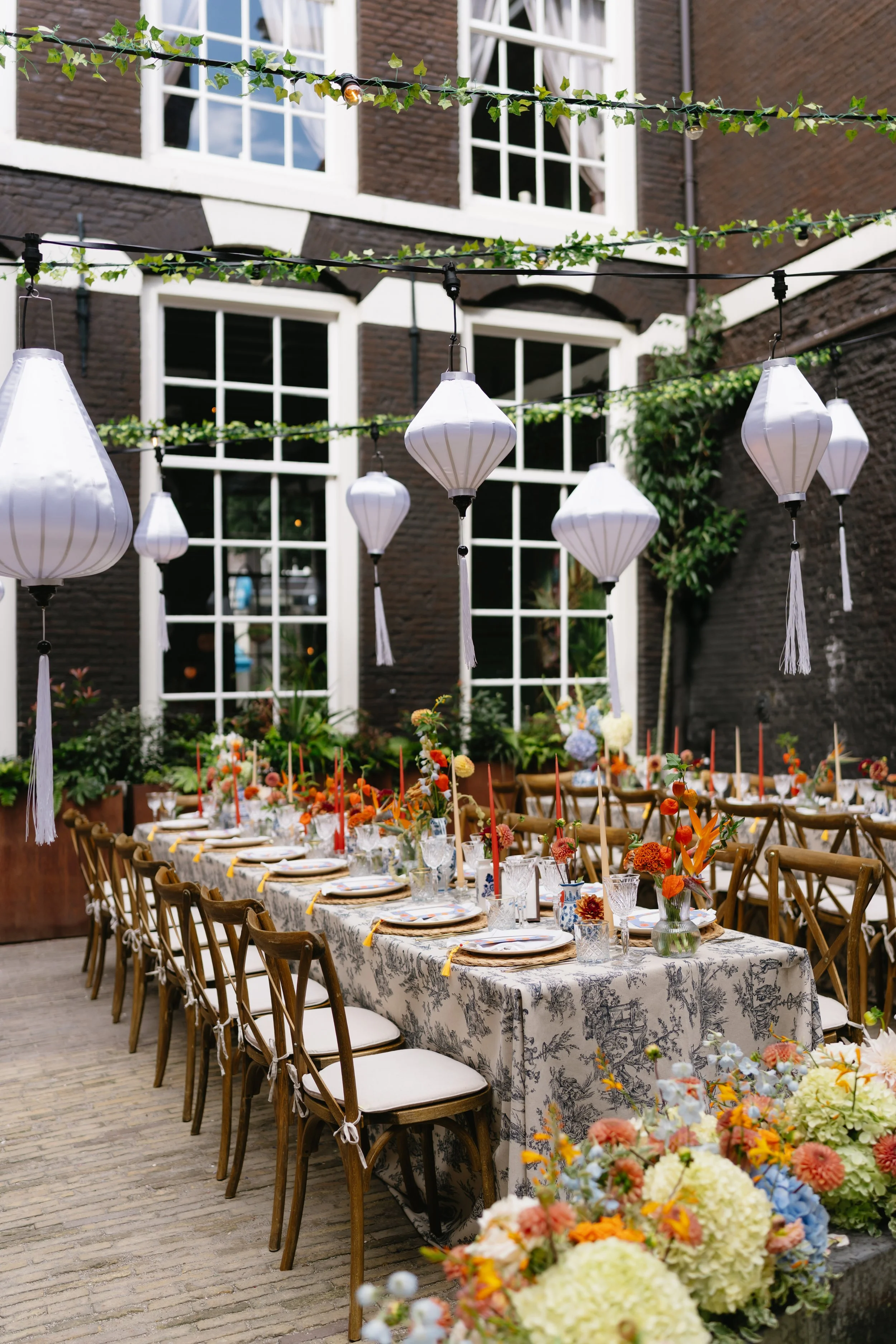 A long banquet table set with floral arrangements and tableware in an outdoor courtyard decorated with hanging lanterns and string lights, with large white-framed windows on a brick building in the background.