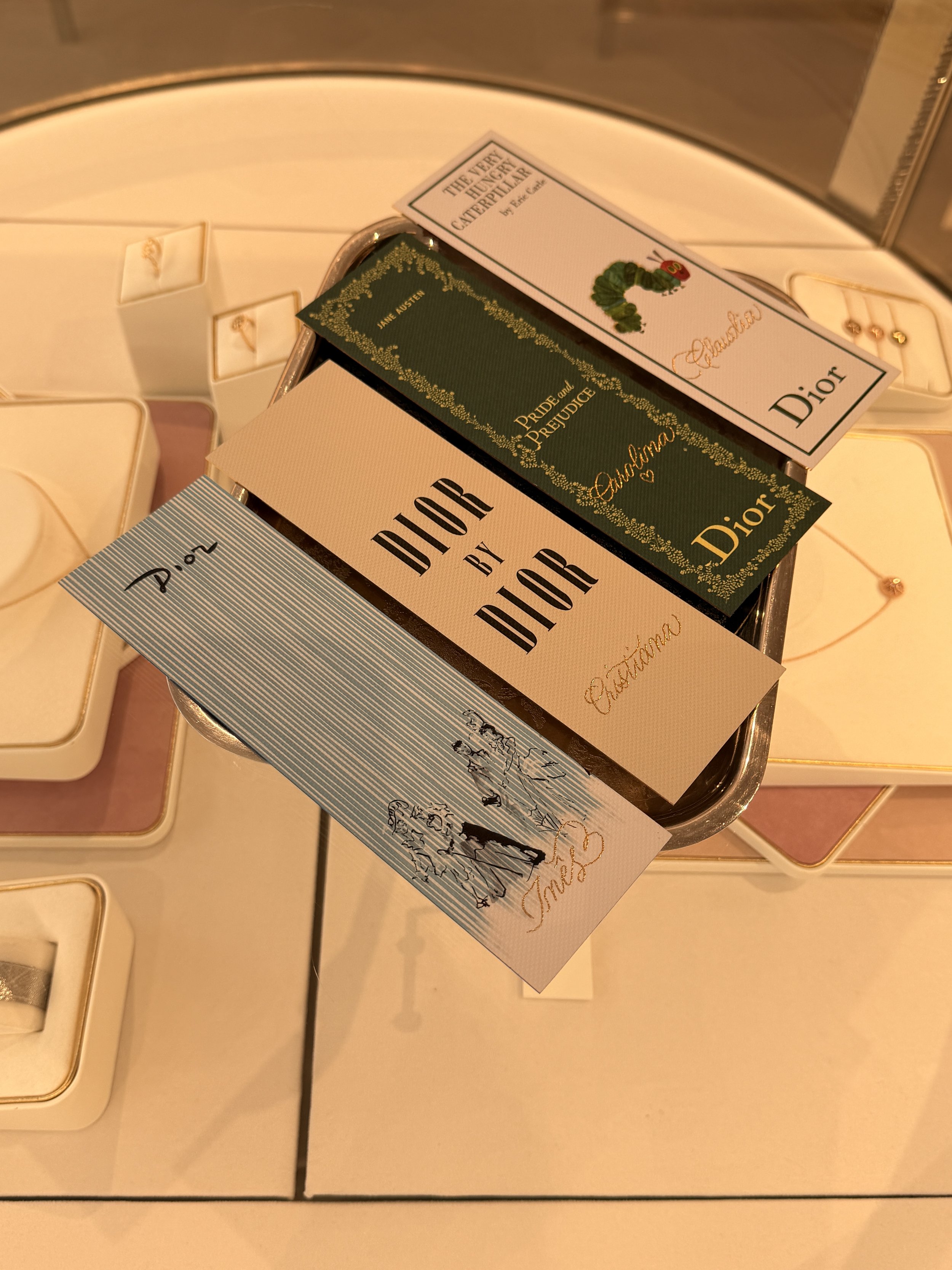 Close-up of three Dior gift boxes, one with a book titled 'The Very Hungry Caterpillar' and another with a card titled 'Pride and Prejudice' surrounded by jewelry on a cream-colored surface.