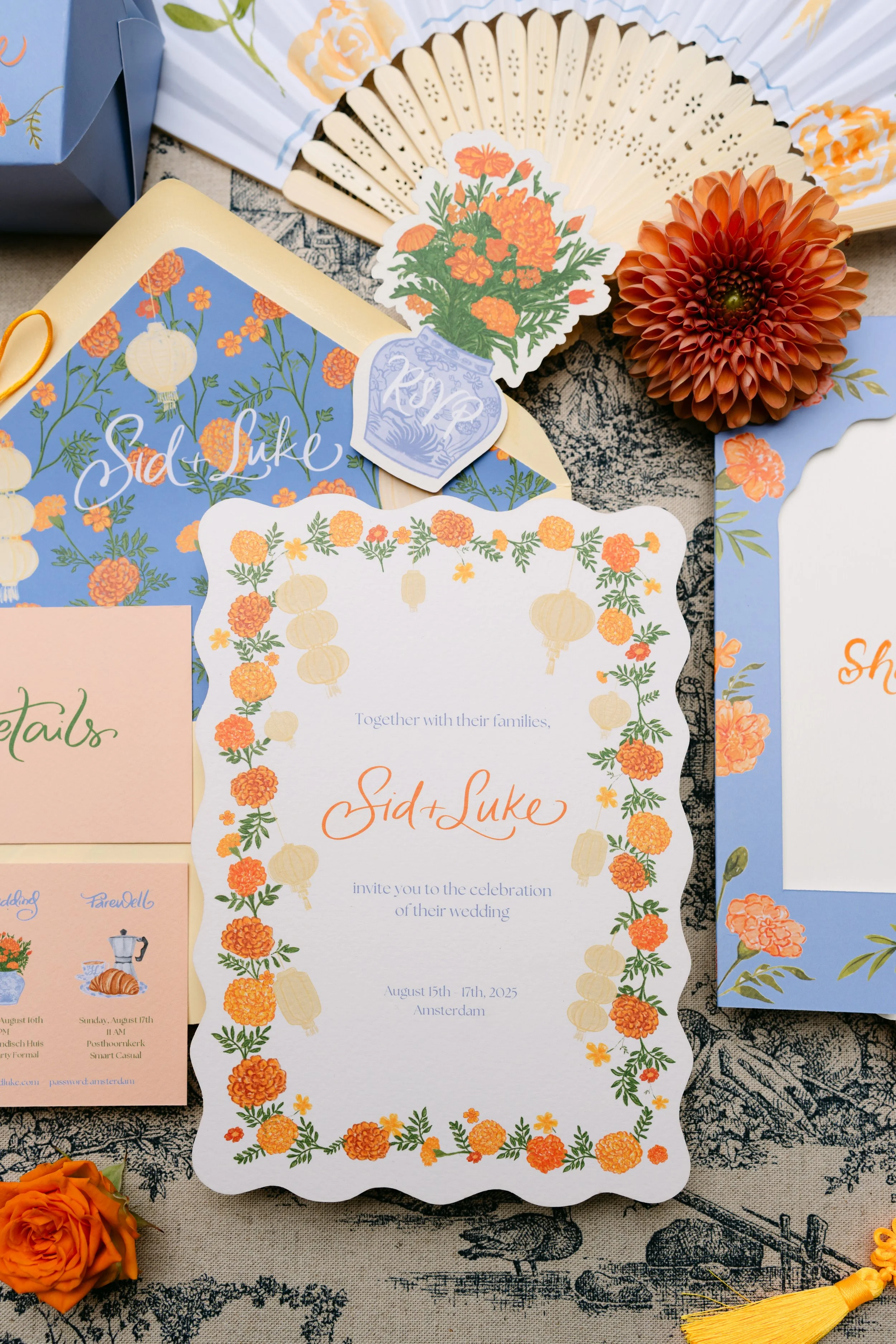 Wedding invitation card with orange and yellow marigold flower border and with text inviting to the wedding of Sid and Luke on August 15th-17th, 2025 in Amsterdam. The card is decorated with floral illustrations and lantern motifs.