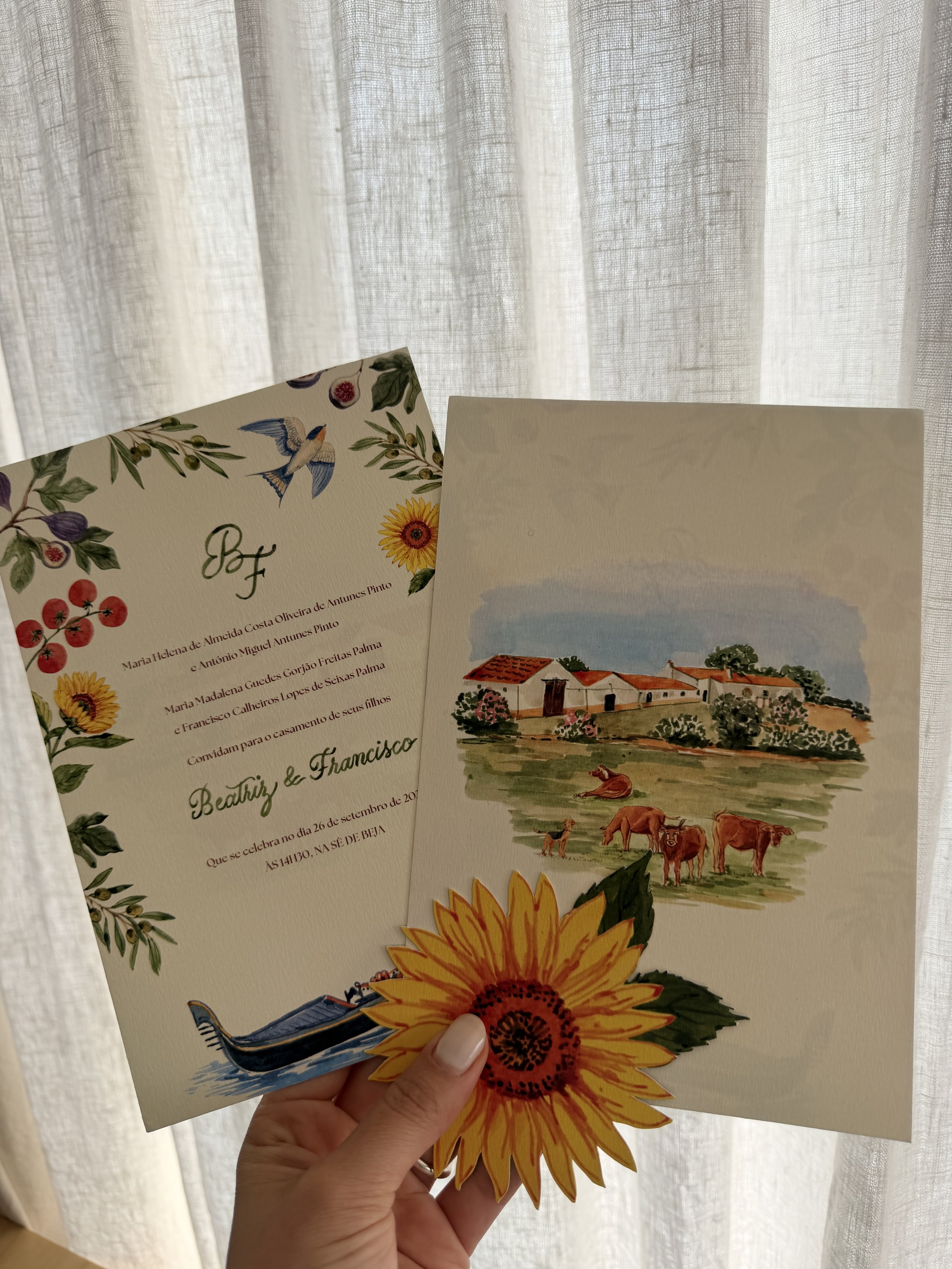 A person holding wedding invitations and a greeting card, with a sunflower decoration at the bottom. The invitations have colorful illustrations of a house, cows, and sky, while the greeting card has a floral and bird design. Portuguese wedding