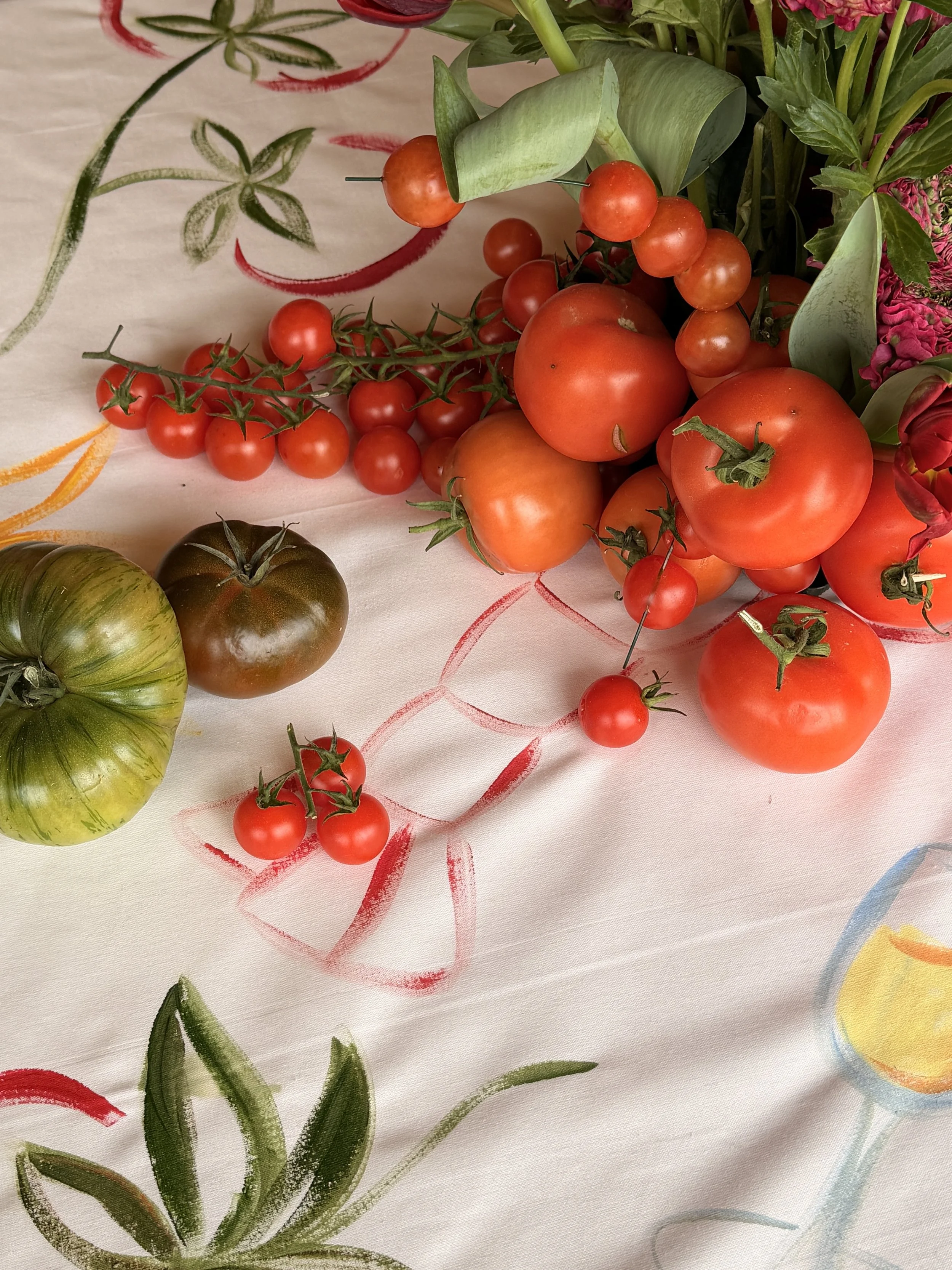 A bunch of cherry tomatoes, some large round tomatoes, green leaves, and a few purple flowers on a tablecloth with colorful decorative patterns.