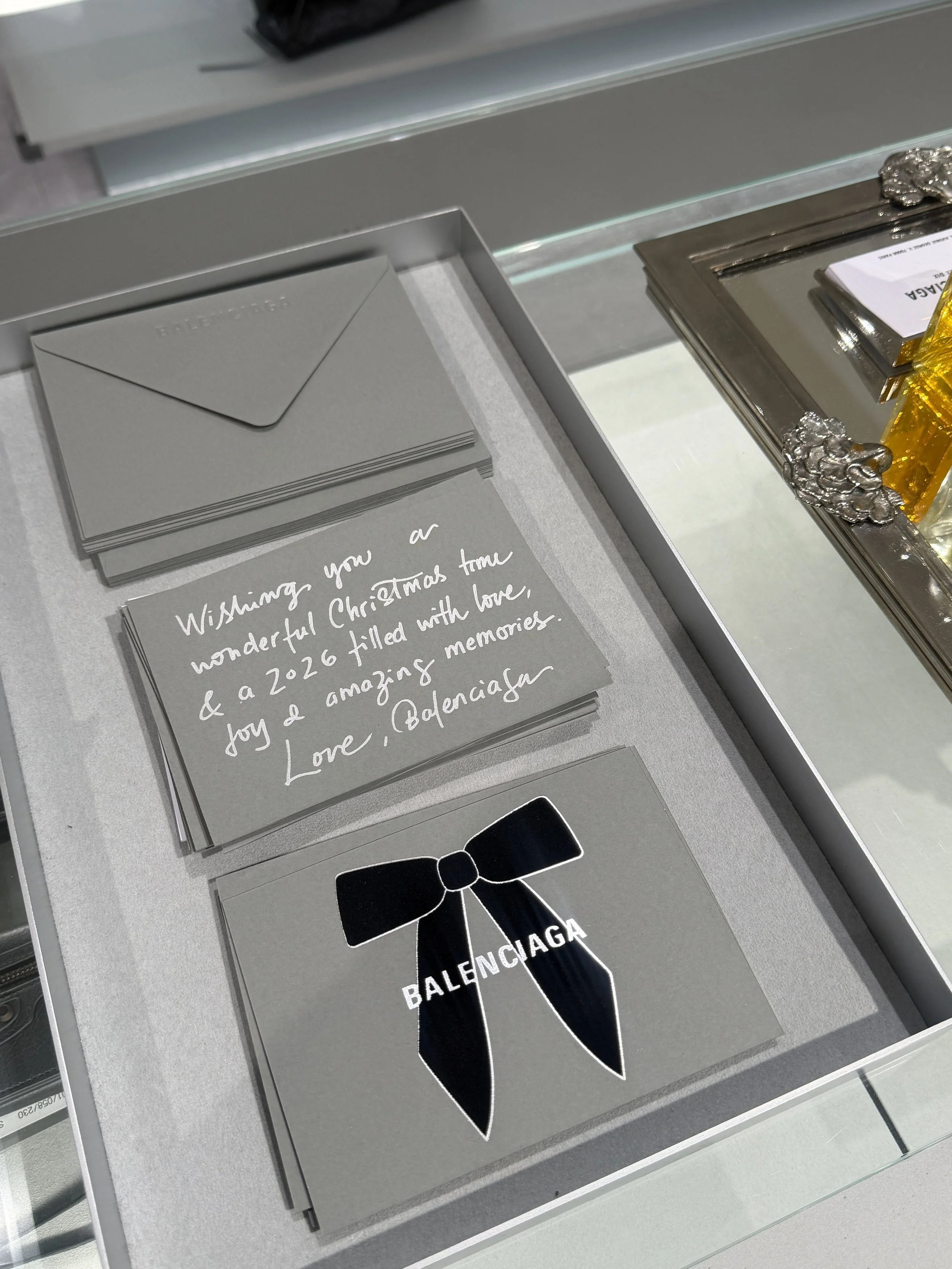 A display case featuring various fashion and holiday themed items, including a card with handwritten Christmas wishes, a black Balenciaga logo ribbon, and silver decorative accents.