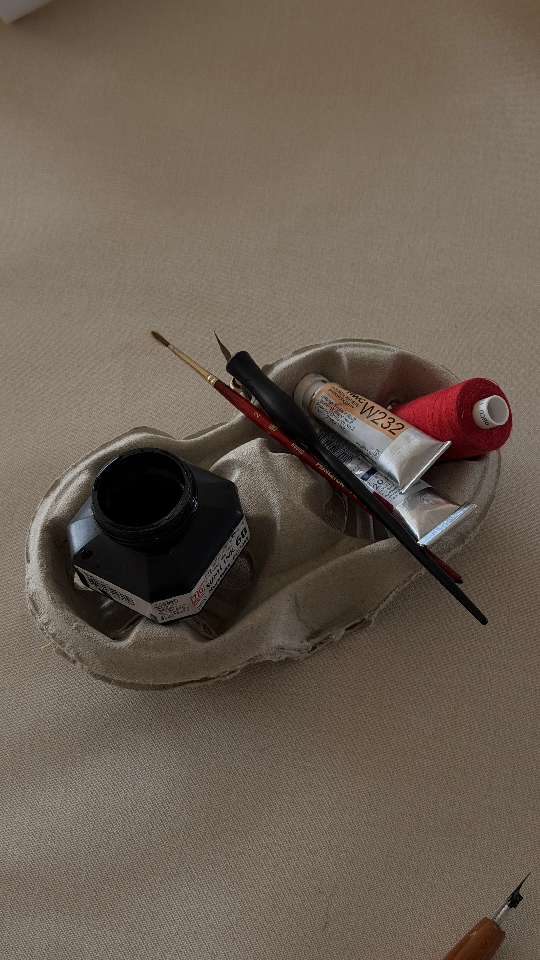 A cardboard egg carton containing a bottle of black ink, red thread, a tube of paint, a paintbrush, and a needle with orange thread.
