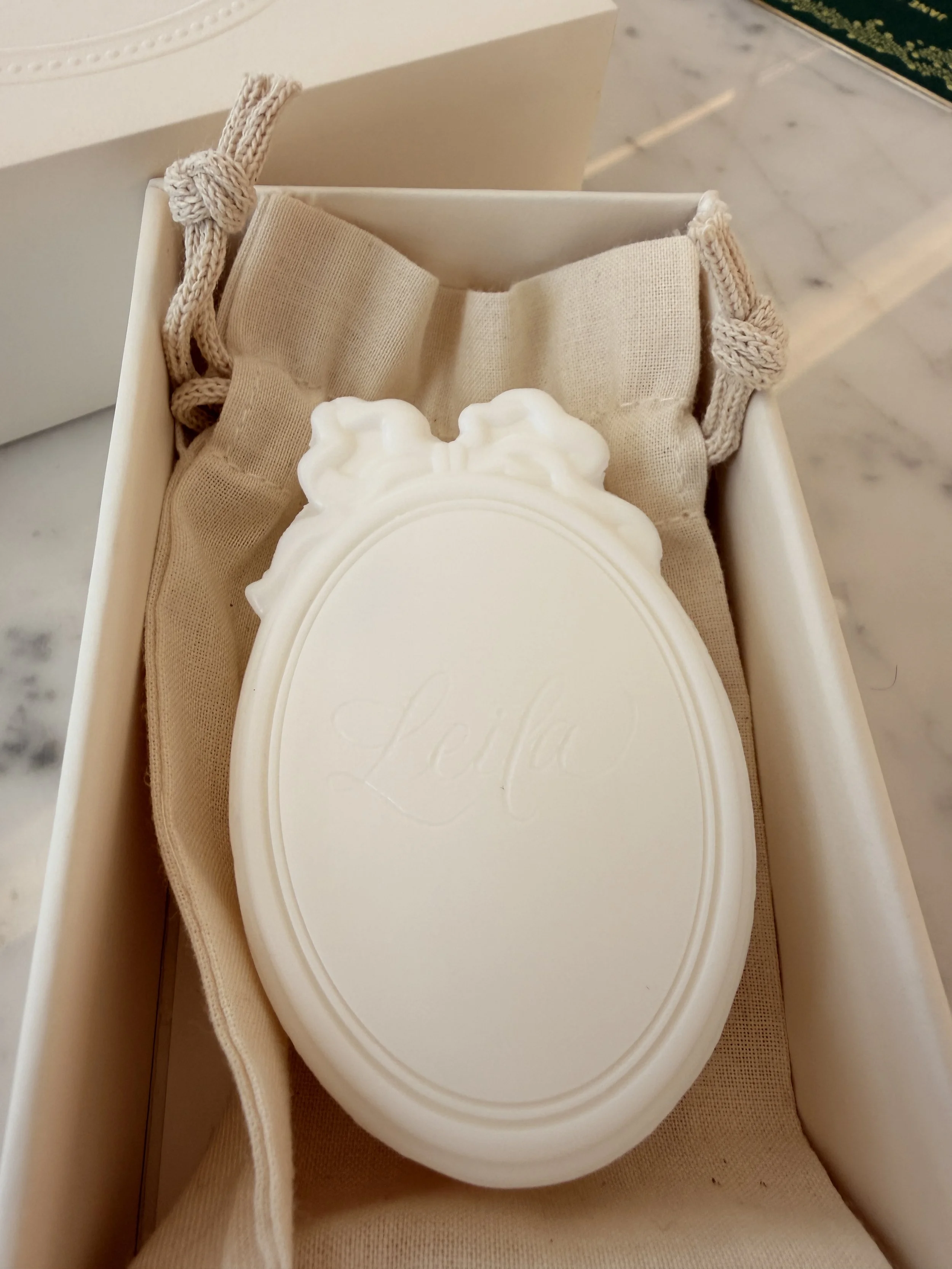 A white decorative soap with the word 'Love' engraved on it, placed on a beige cloth inside a white box.