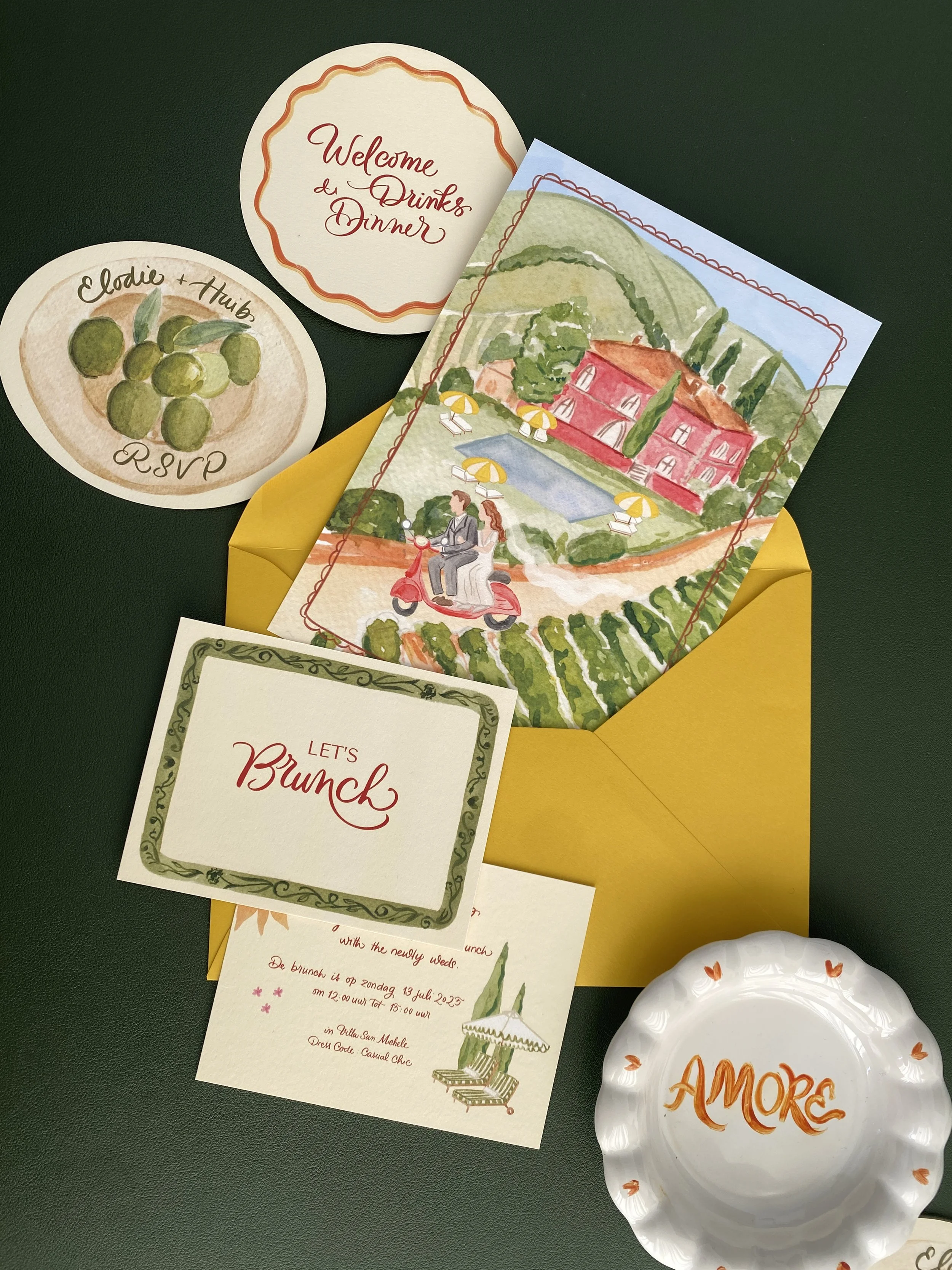 A set of invitation cards and a small dish on a dark green surface. The cards include a round 'Welcome & Drinks Dinner' sign, a 'RSVP' card with a picture of green grapes, a large illustrated card of a house with a pool and a couple on a scooter, and