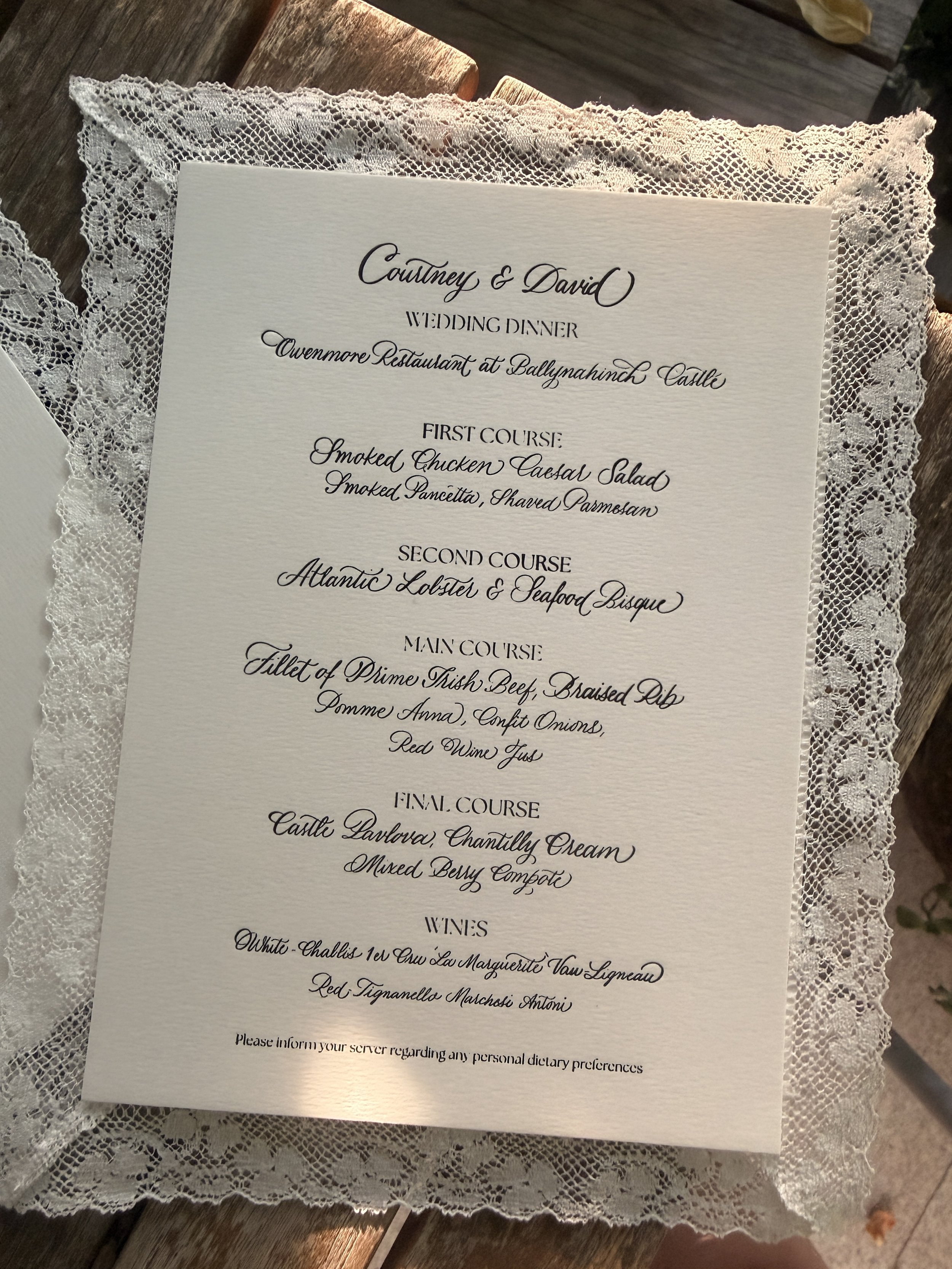 A wedding menu printed on textured paper with elegant cursive font, placed on a lace-edged cloth on a wooden surface. For Olivia Buckley International. 