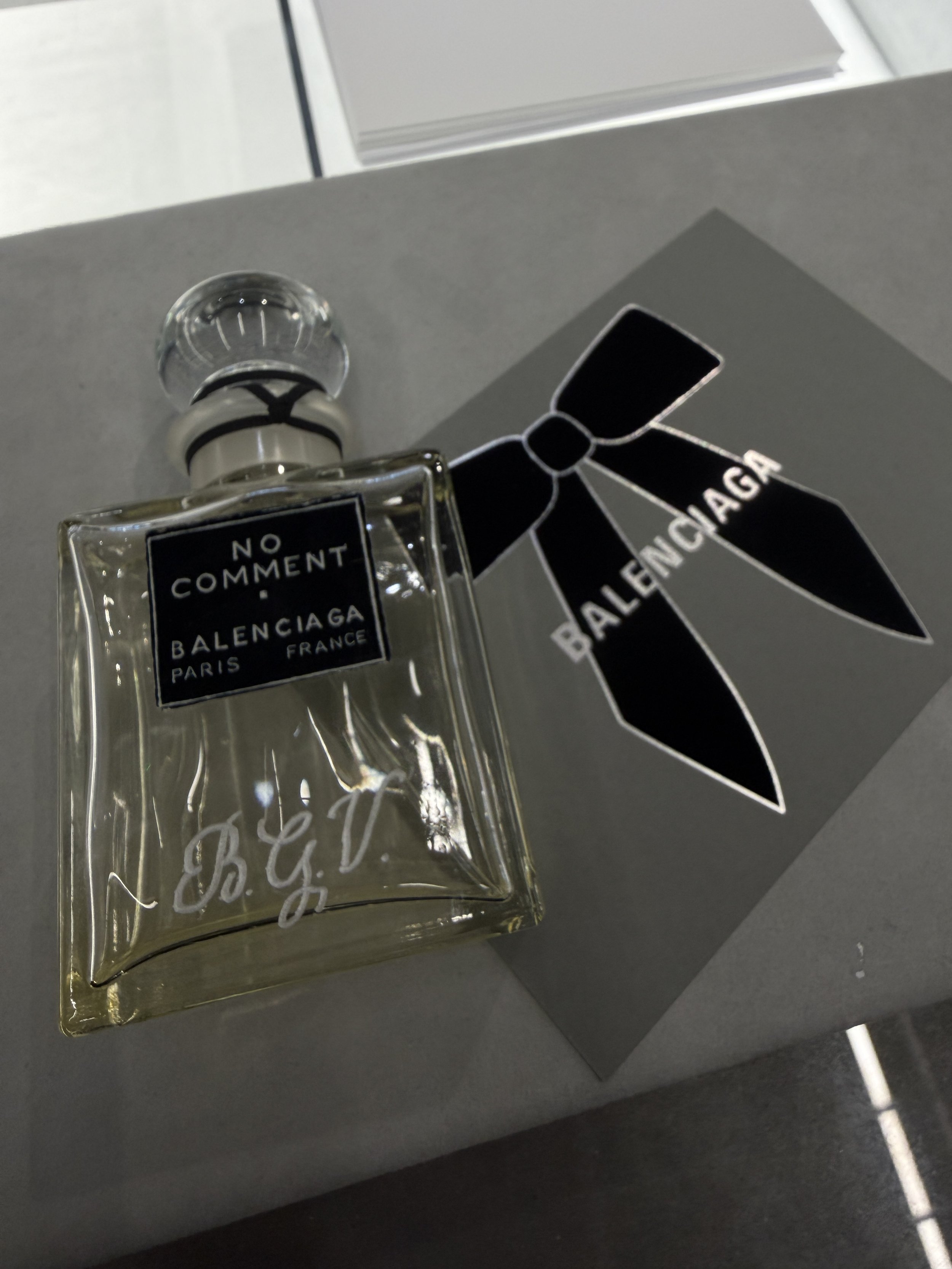 A clear glass perfume bottle with a black and white Balenciaga label, a black ribbon bow, and decorative black and white tag, placed on a dark surface.