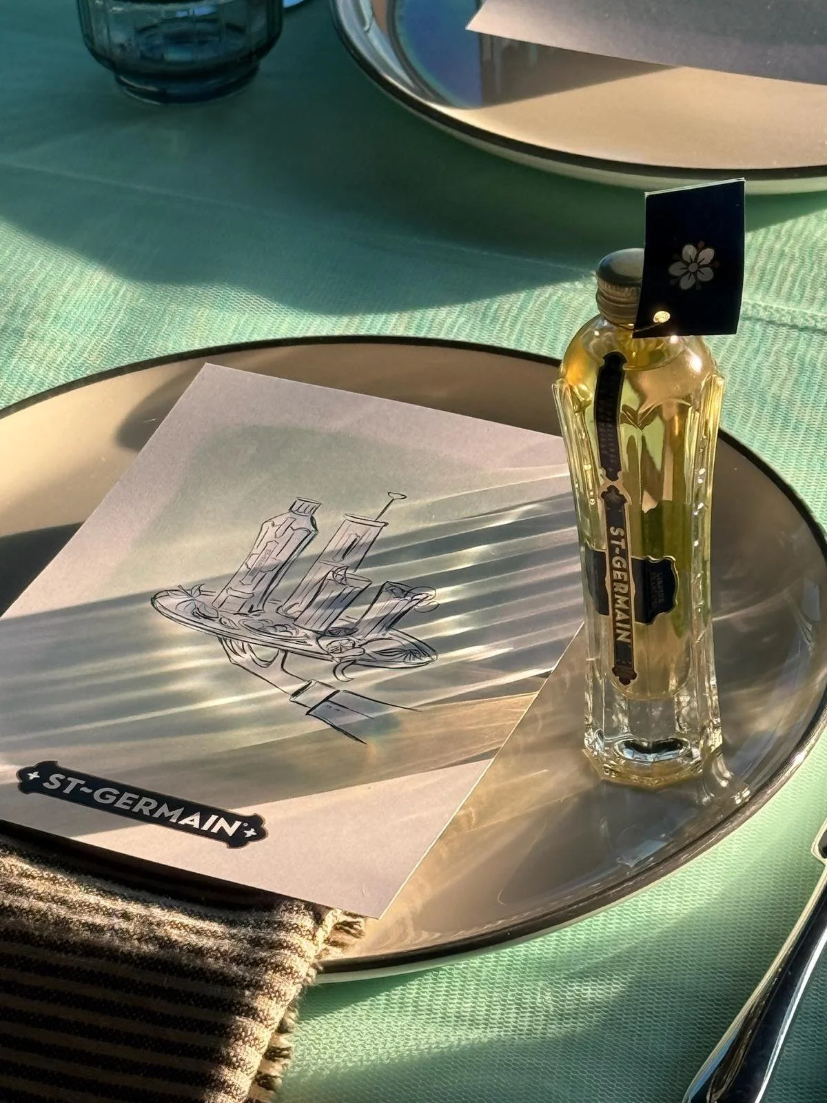 Illustrated menu displayed on a silver plate beside a St-Germain bottle at a green table setting