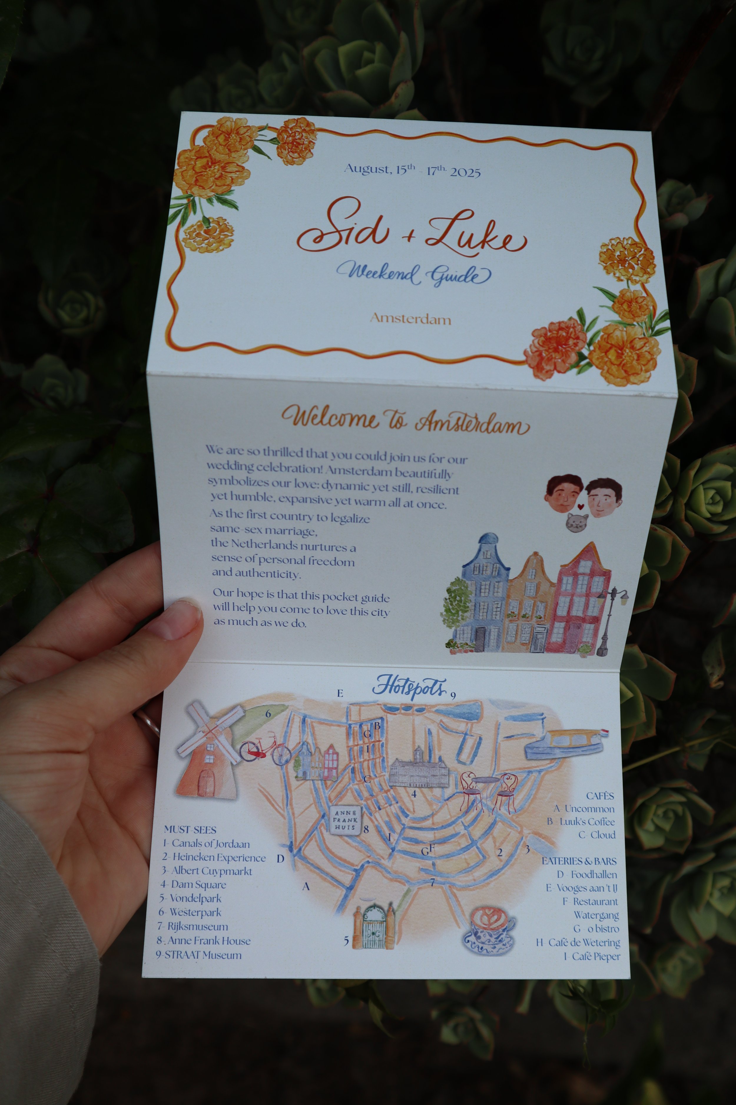 A hand holding a folded travel guide map of Amsterdam, Netherlands, with illustrations of buildings and a map of attractions inside, set against a background of green succulent plants.