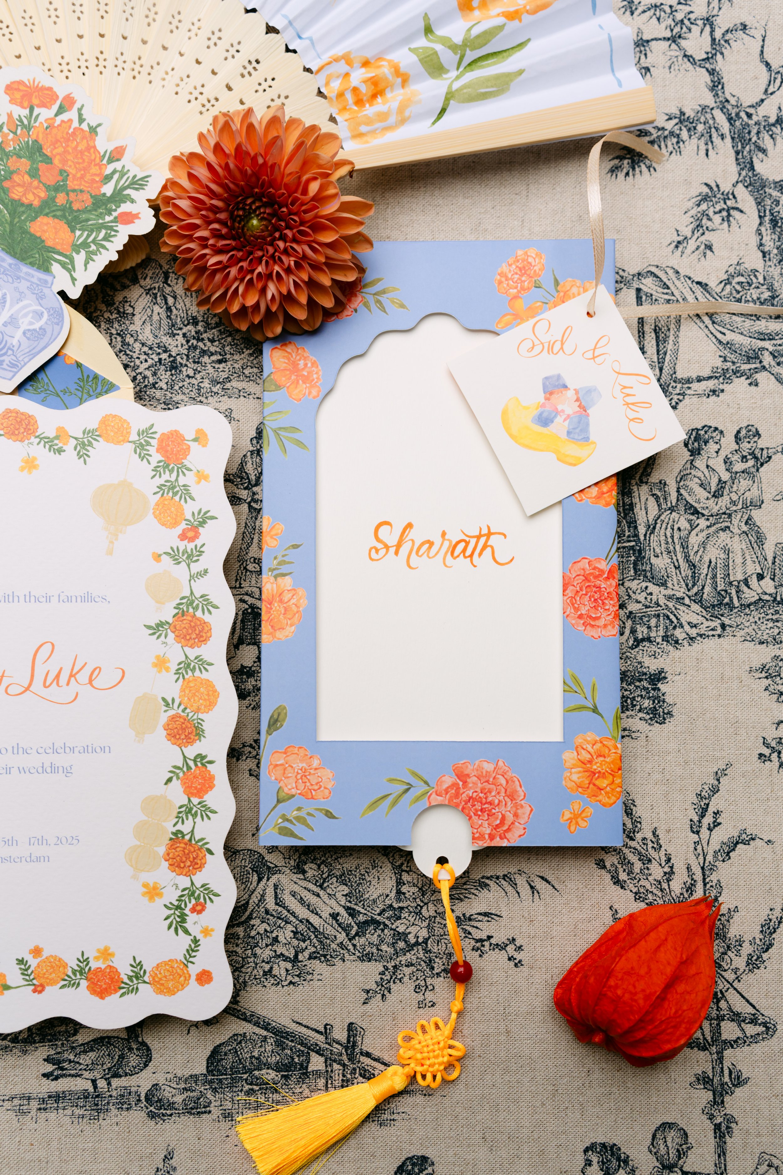 A floral-themed photo card with the name 'Sharath' on it, decorated with orange flowers, and a tag with 'Sid & Lulo' and a drawing of colorful blocks and a banana. There are paper cutouts with orange flowers, a pom-pom string with a yellow tassel, an