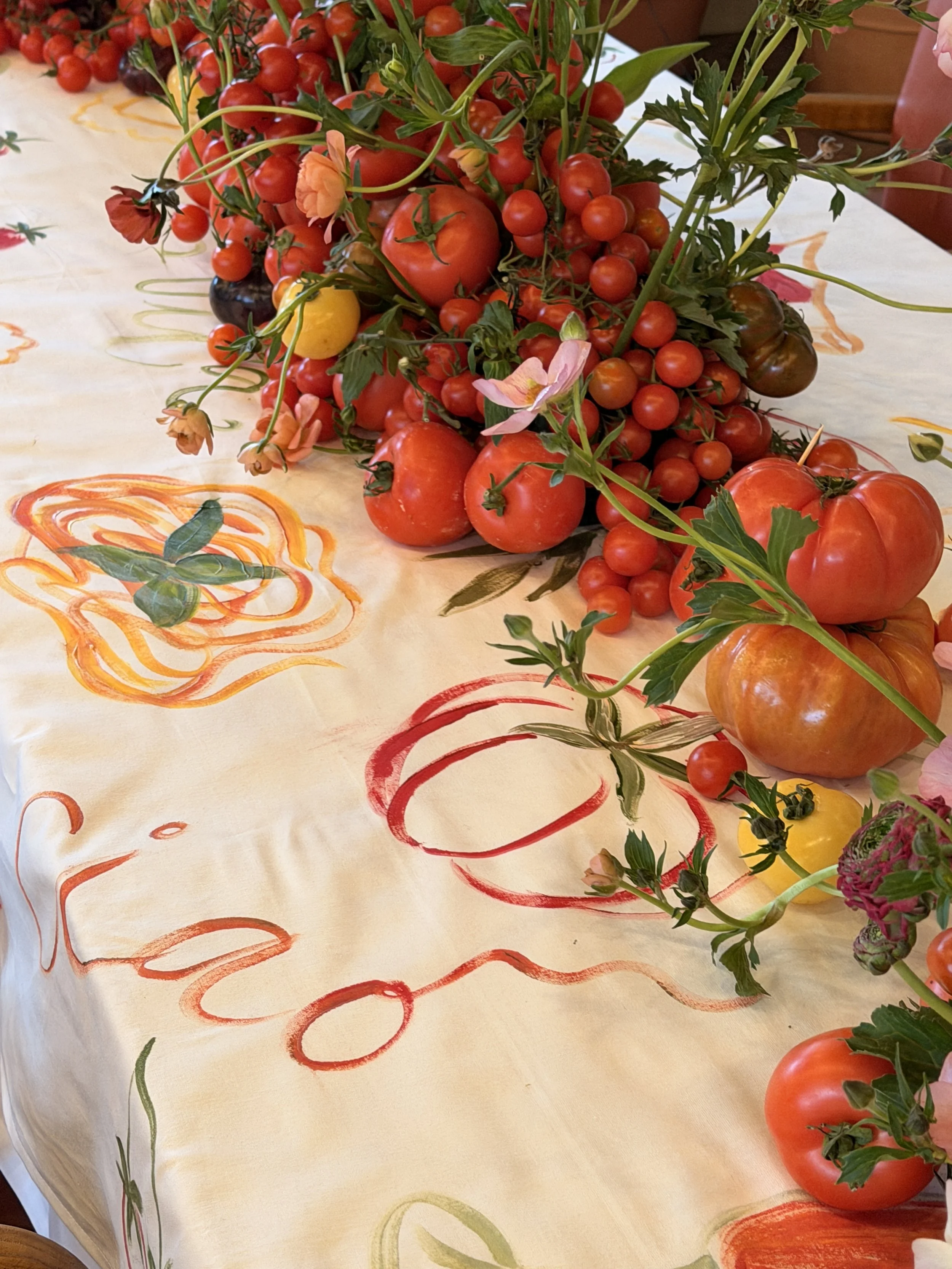 A table with a floral tablecloth decorated with an arrangement of various ripe tomatoes, including red, yellow, and black varieties, with some pink flowers and green leaves.