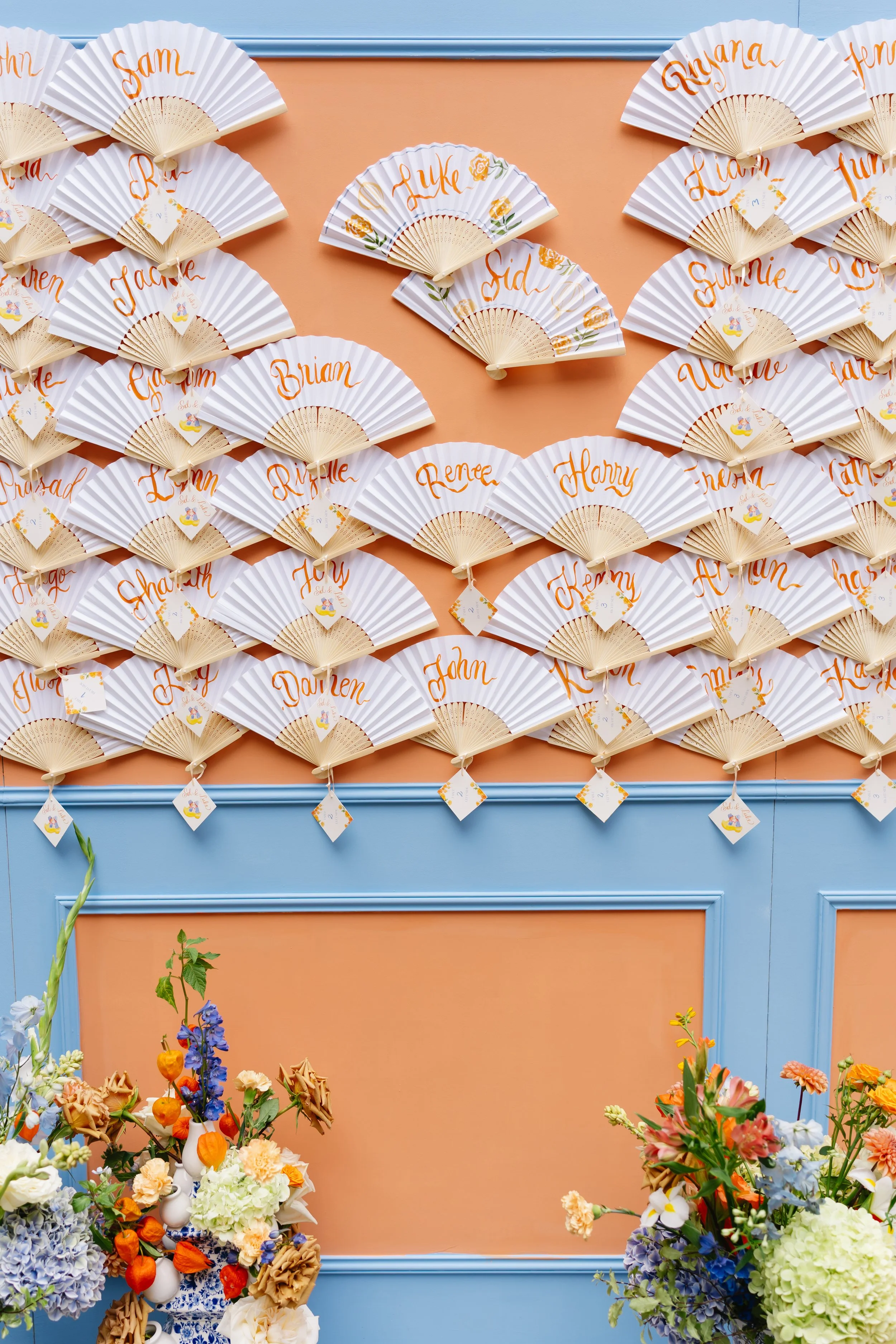 Display of personalized paper hand fans with names written in orange, arranged on a peach-colored wall with blue framing, and decorated with fresh floral arrangements in front.