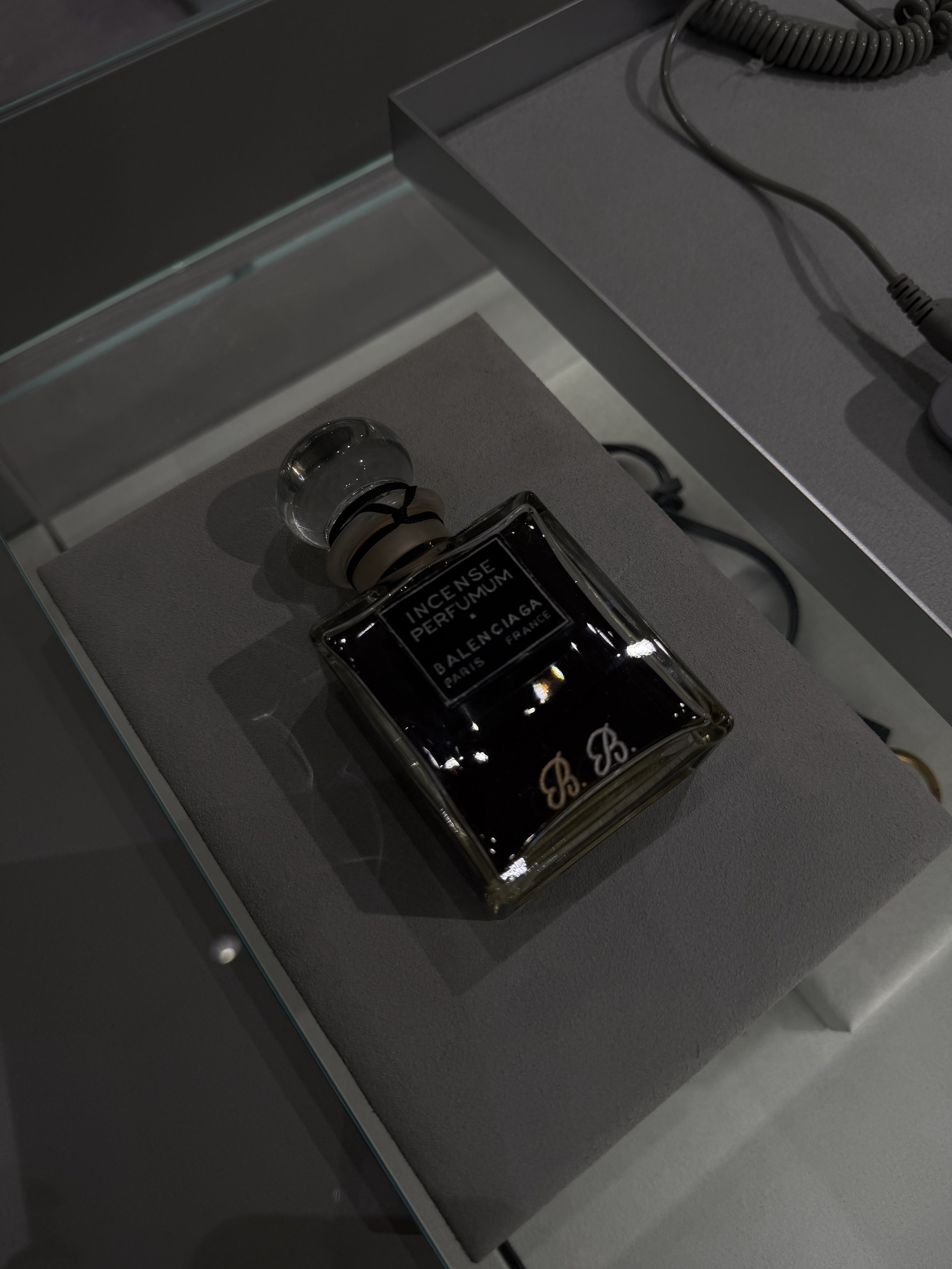 Close-up of a rectangular perfume bottle labeled 'Incense Perfume' from Balenciaga, with a black liquid inside, placed on a display surface with a coiled electrical cord nearby.