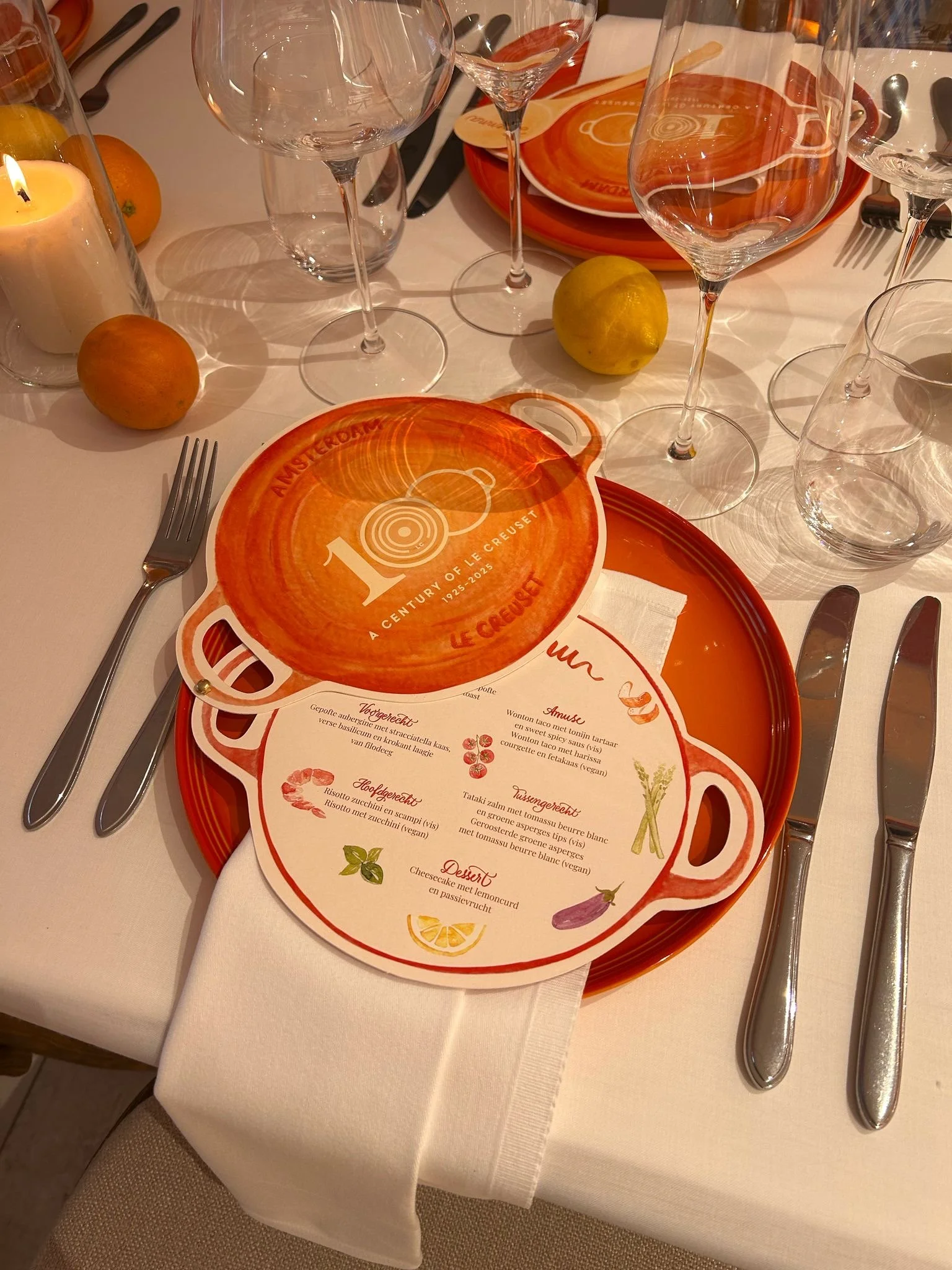 A table setting with a menu, orange plates, glasses, cutlery, a candle, an orange and a lemon, and a napkin on a white tablecloth. The menu is decorated with illustrations of vegetables and has a logo that celebrates 100 years of Le Creuset.