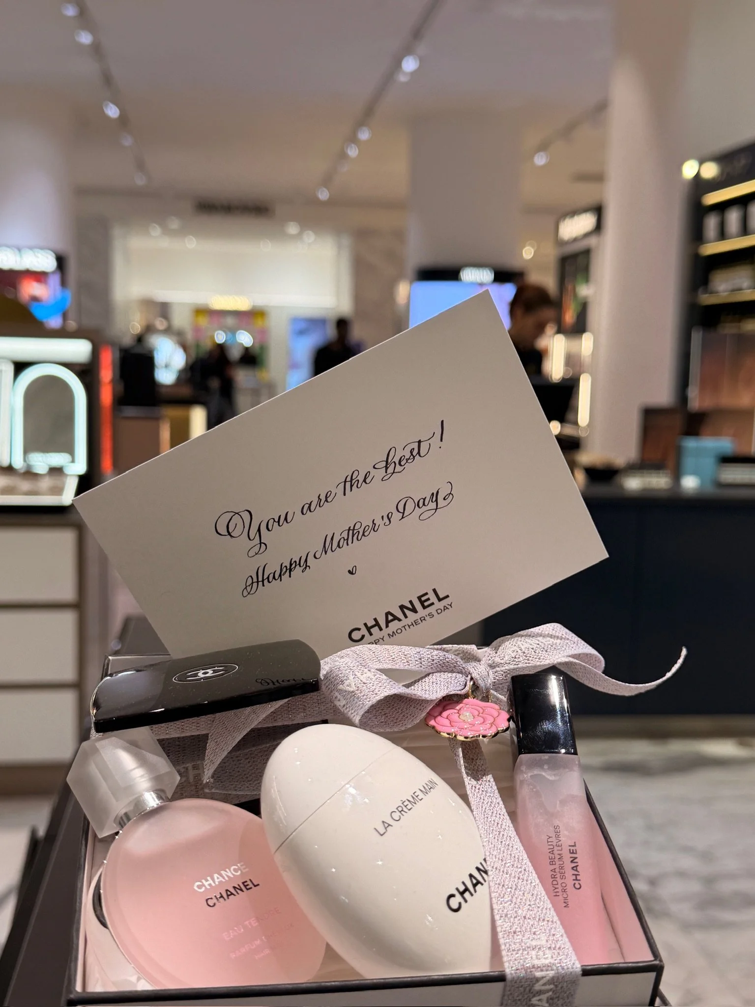 A collection of Chanel beauty products, including Chance Eau Tendre perfume, La Creme Main hand cream, and Hydra Beauty micro serum, arranged in a box with a pink flower charm and a white ribbon, with a shopping mall background.