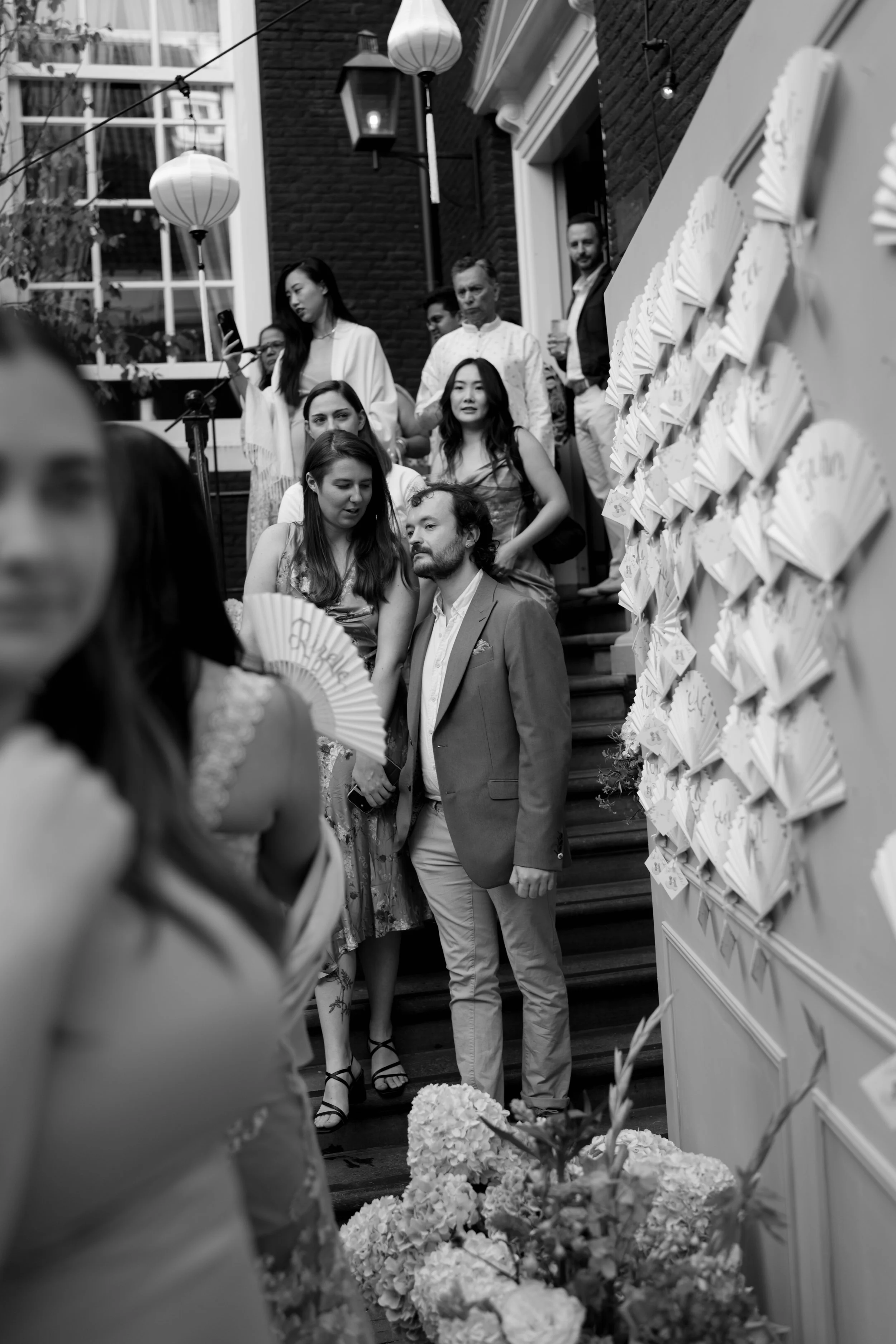 Guests at an indoor event gathered on a staircase with floral decorations, some holding drinks and some appearing to engage in conversation.