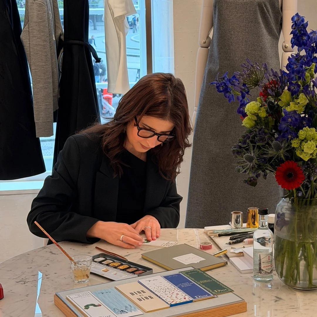 A woman with dark brown hair, glasses, and a black blazer is sitting at a marble table, working on a project with various art supplies and paper in front of her. There is a large vase of colorful flowers on the table and clothing on mannequins in the background.