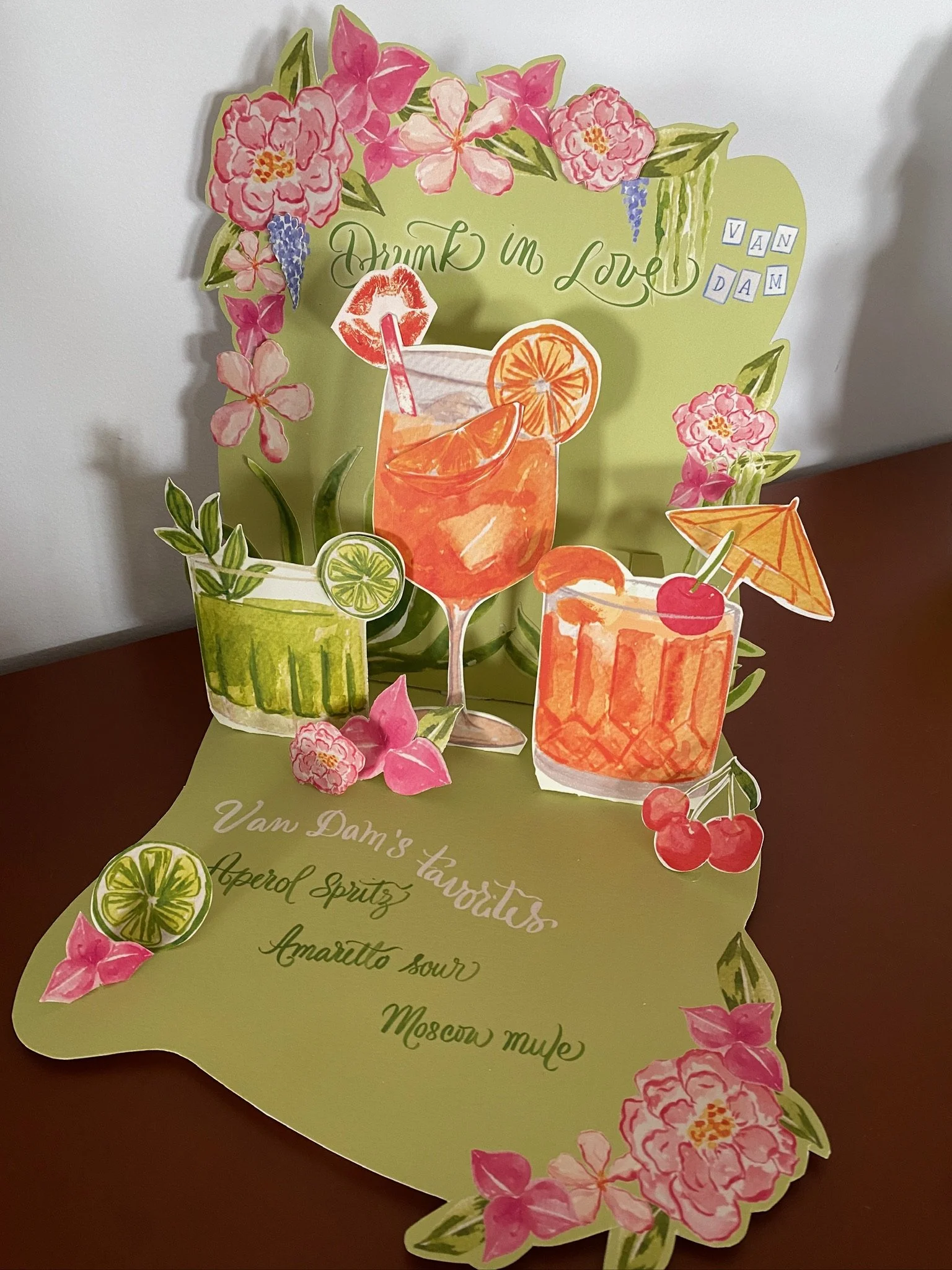 A colorful paper cutout display 3D design featuring illustrations of various cocktails with garnishes such as fruit slices and cherries, decorated with pink flowers and green leaves, and the phrase "Drink in Love" written in green cursive text.