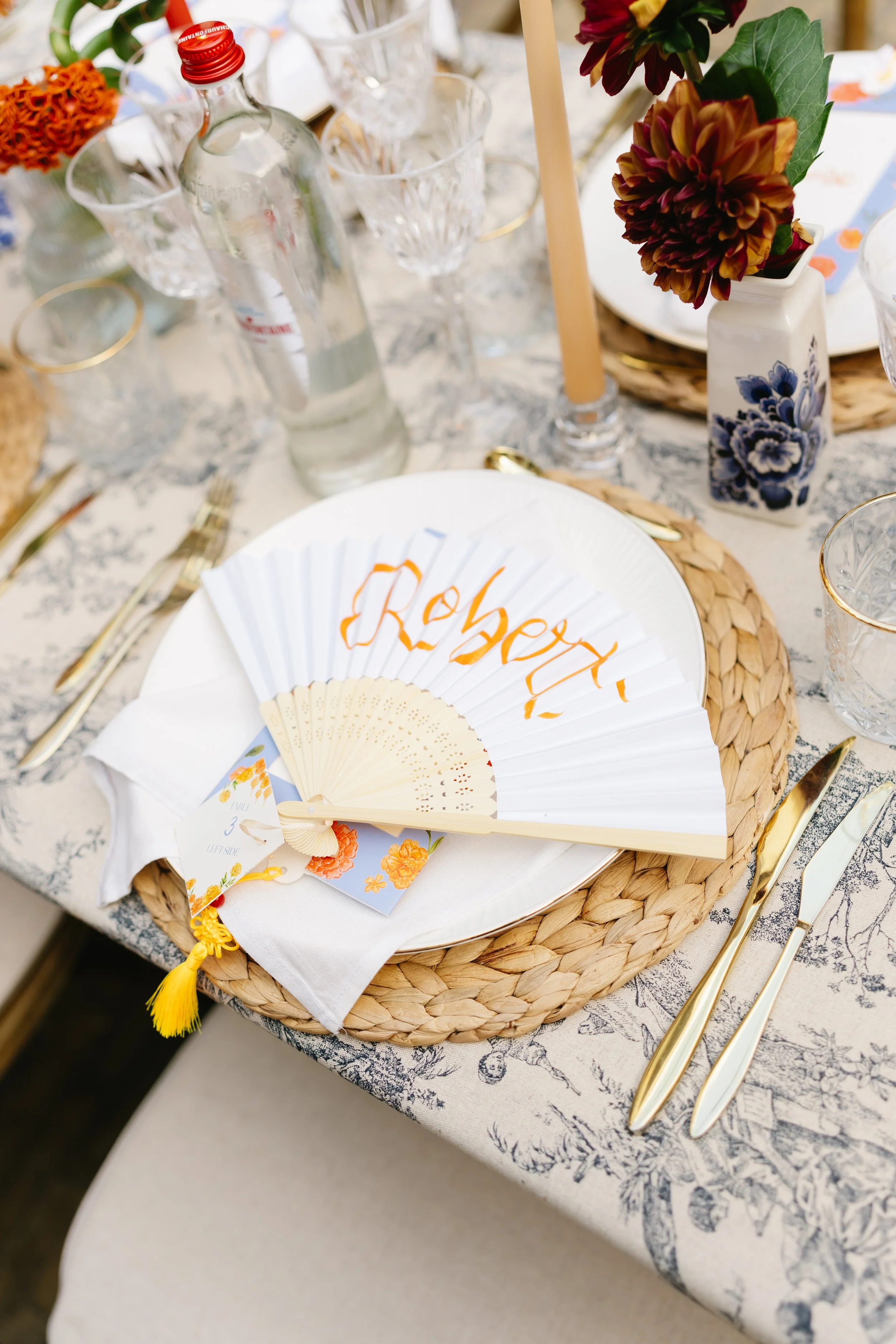 Close-up of an elegantly set table with a white ceramic plate, a cream-colored hand fan with orange writing, gold utensils, a woven placemat, and a centerpiece of flowers in a blue and white vase. A clear water bottle, a candle, and glassware are als
