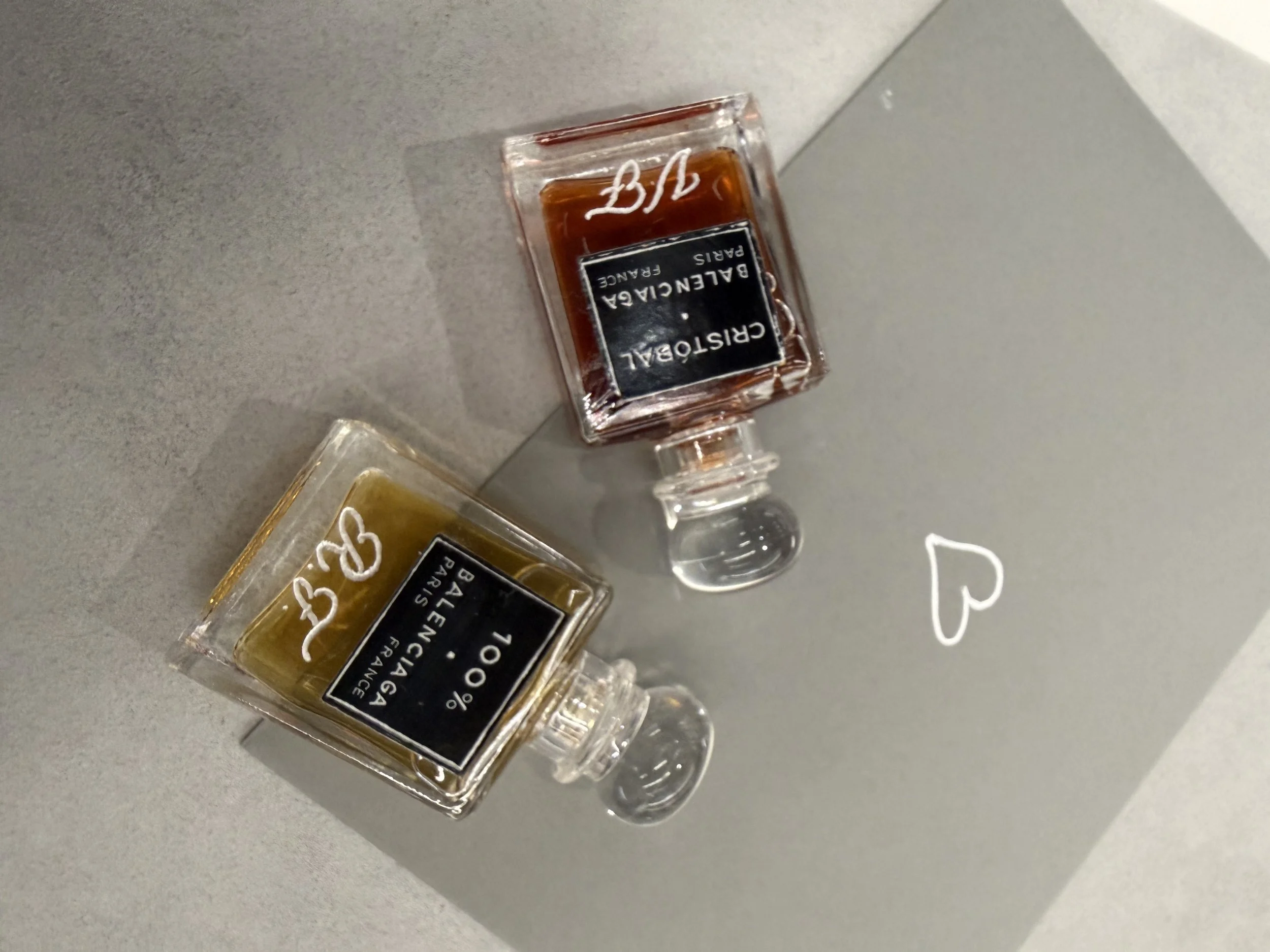 Close-up of a small bottle of perfume labeled 'Balenciaga Paris' lying on its side on a grey surface, with a small clear glass stopper next to it. Hand Engraving by Gaia Amsterdam