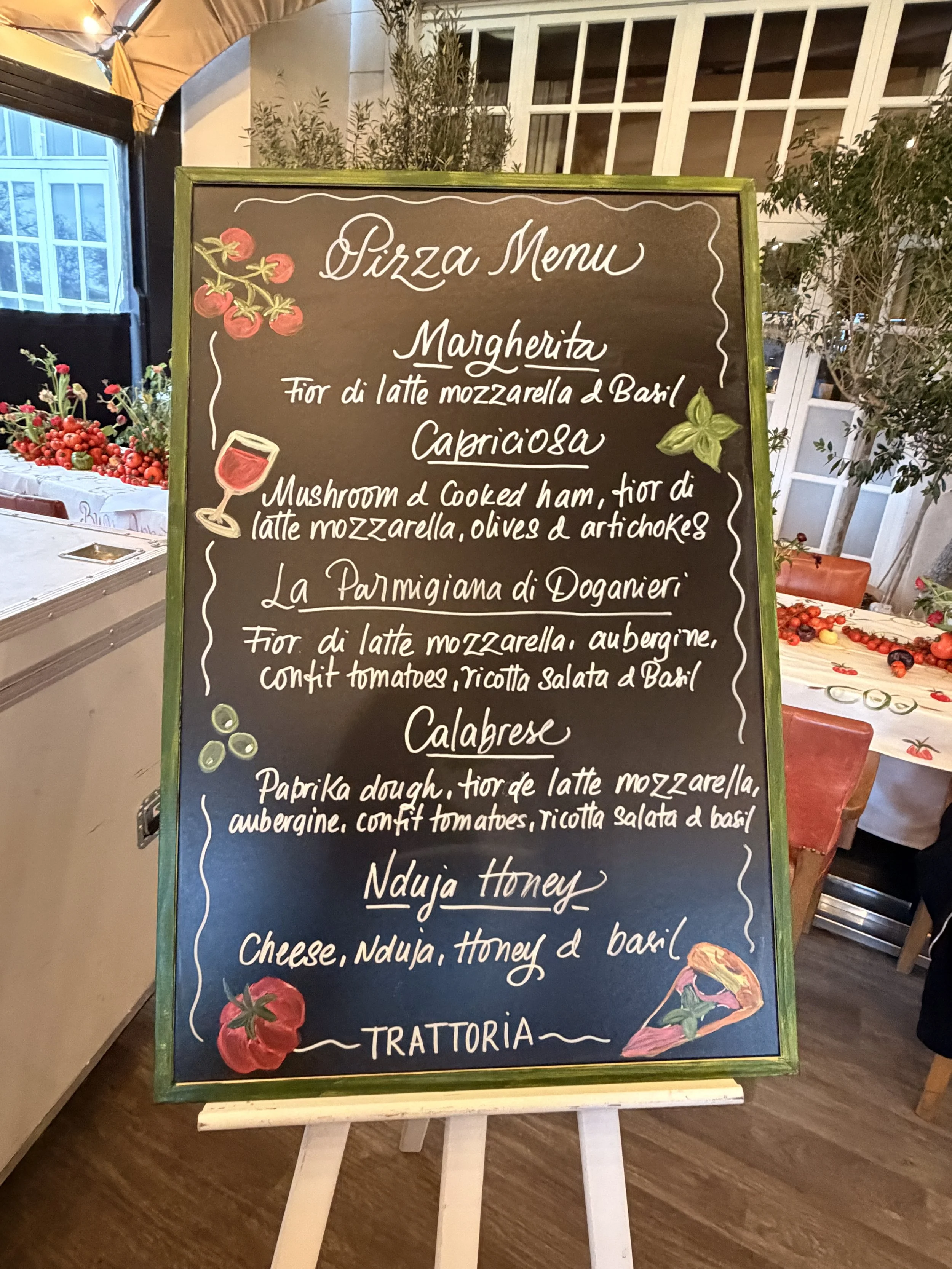 A blackboard menu with colorful illustrations of tomatoes, basil, a glass of wine, and a slice of pizza. The menu lists various types of pizza with their ingredients in Italian and English, including Margherita, Capricciosa, Calabrese, and Nduja Hone