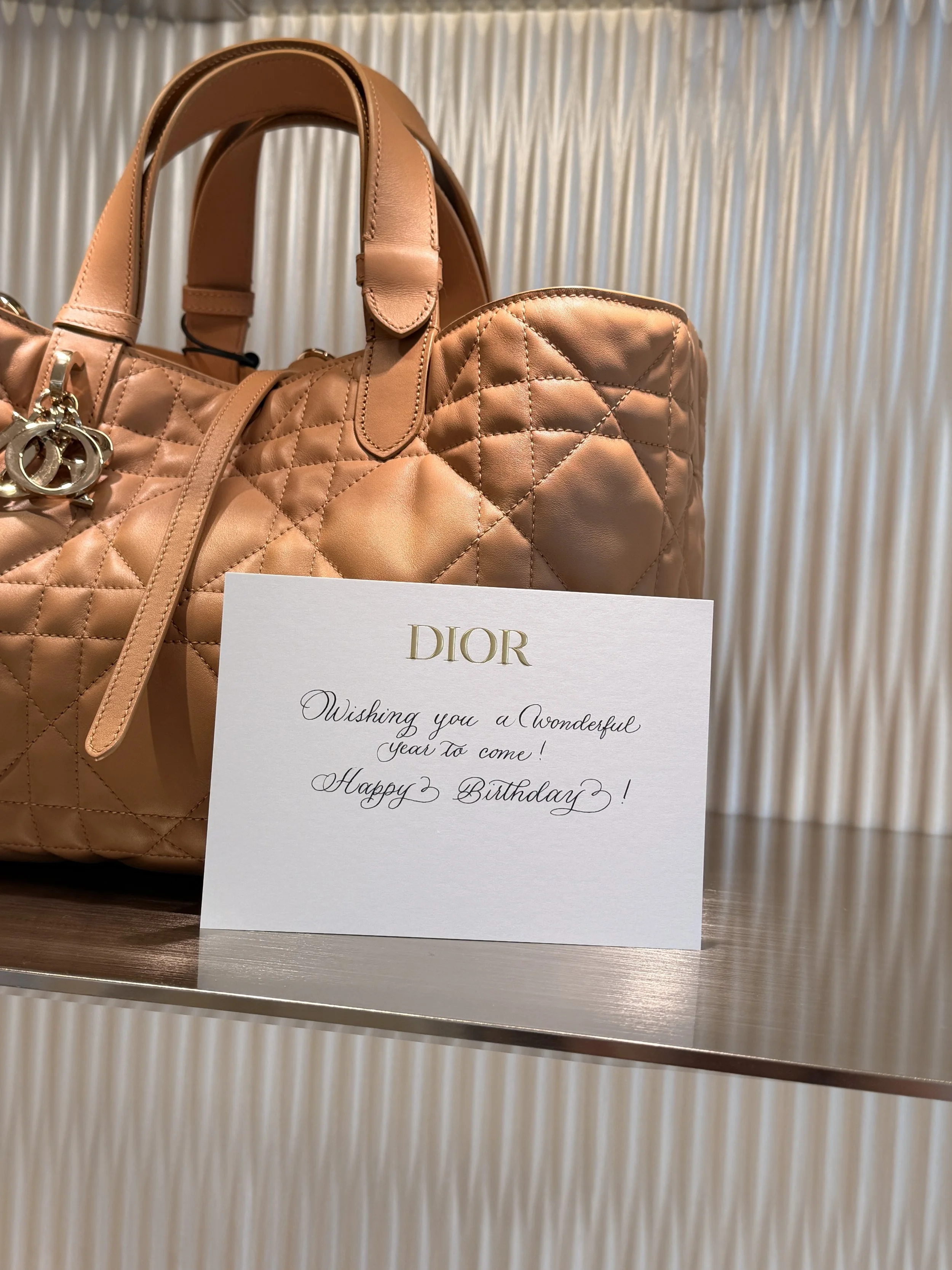 A light brown quilted leather handbag with a Dior charm hanging from the handle, placed on a surface next to a white greeting card with a calligraphy birthday message, against a textured background.