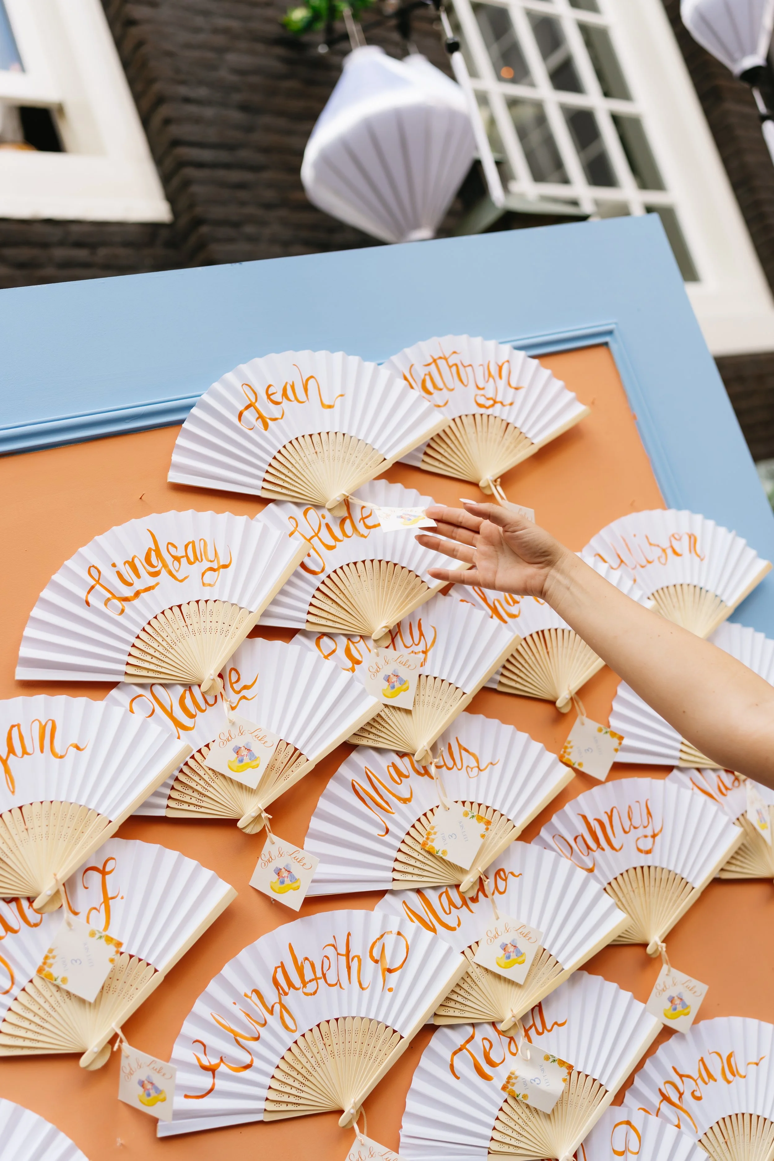 Decorative fans with orange cursive writing, arranged on a peach-colored display board, with a person’s hand adjusting one fan.