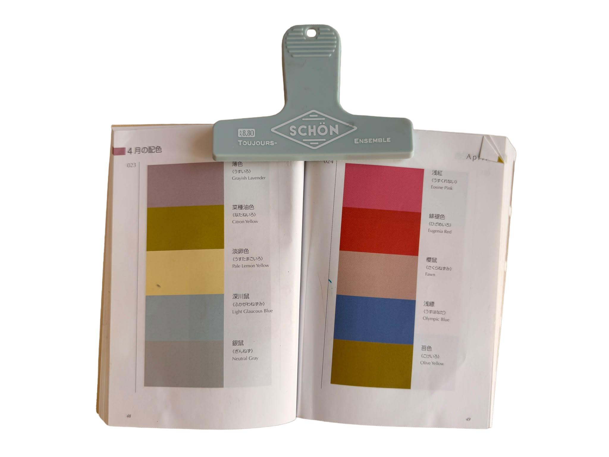 Open color swatch book with various shades of yellow, pink, blue, gray, and green, labeled in multiple languages. A gray stapler is clipped onto the top of the book.