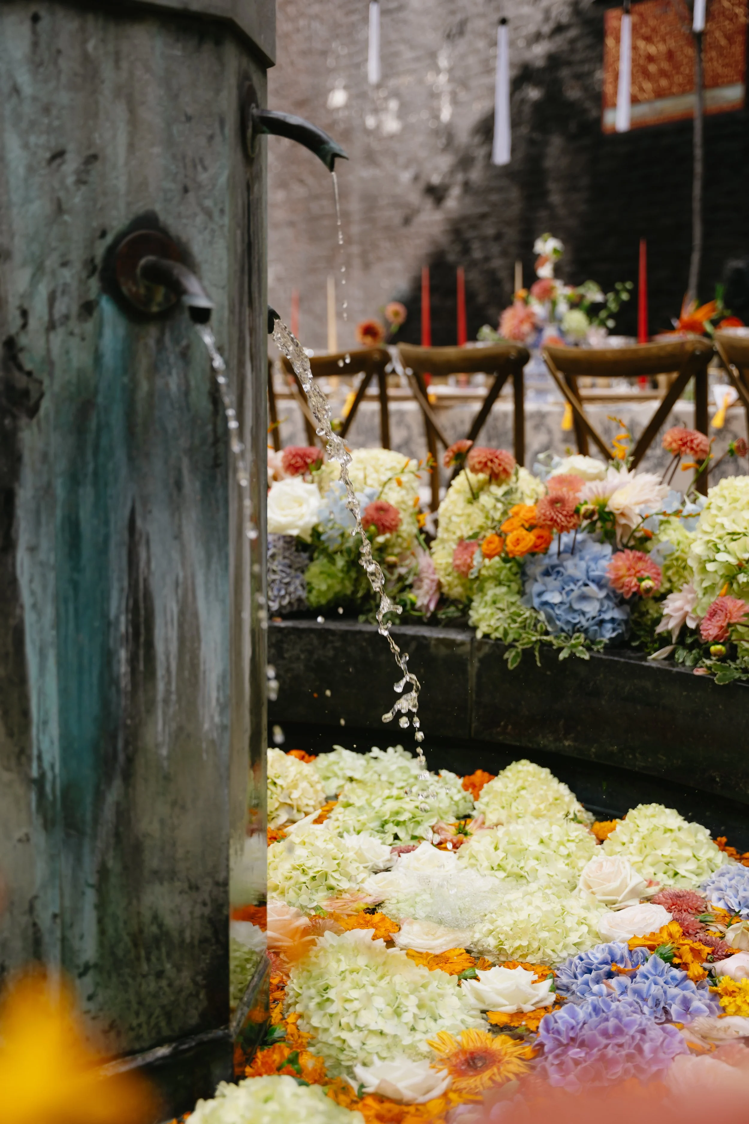 A water spout from an old wooden water fountain, with colorful flower petals and arrangements surrounding it and in the background, possibly at a memorial or celebration event.