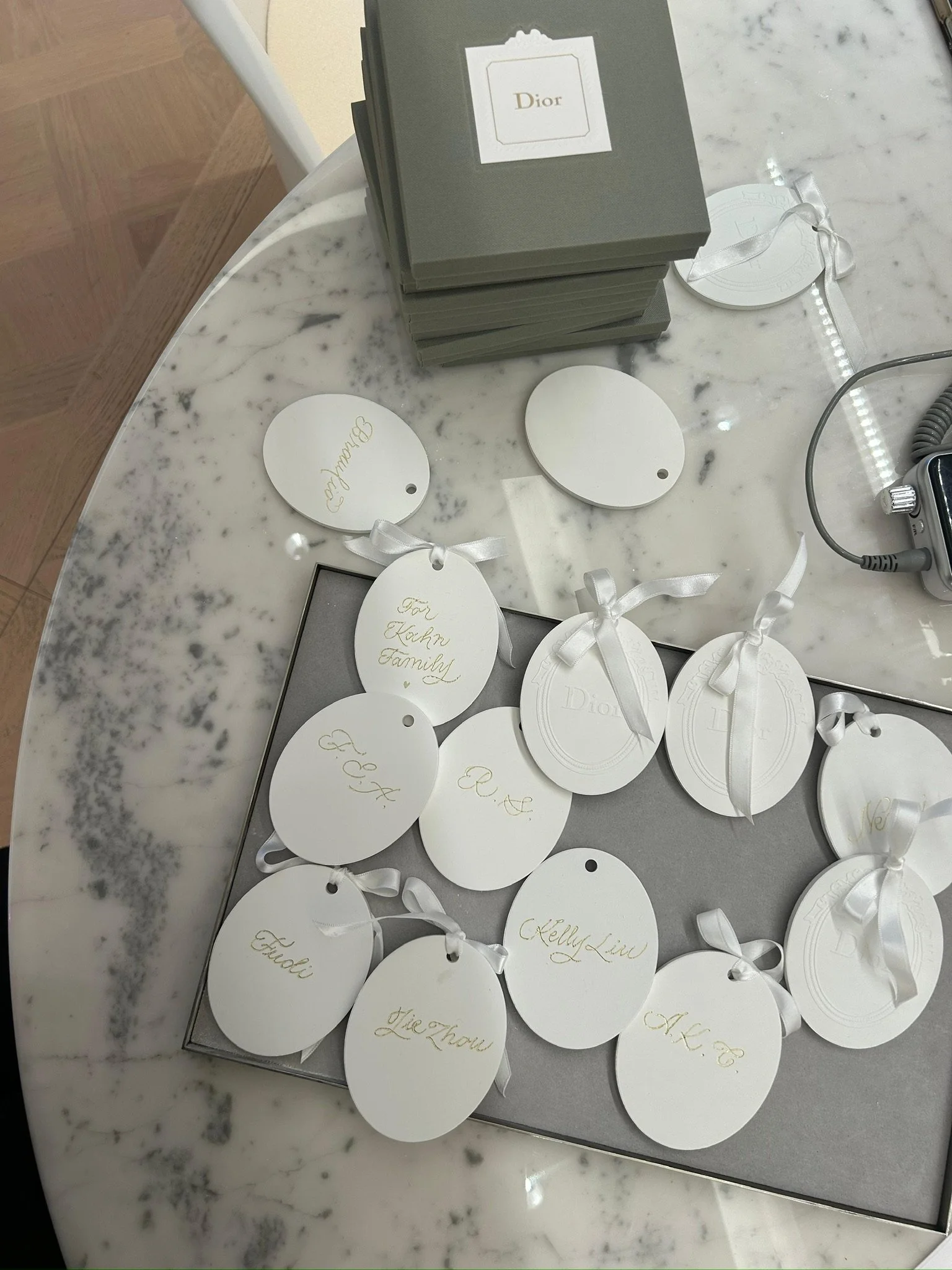 A marble table with a stack of gray paper folders labeled 'Dior', surrounded by white oval name tags with gold script, some with ribbons and some with engraved text, and a silver ribboned seal on top.