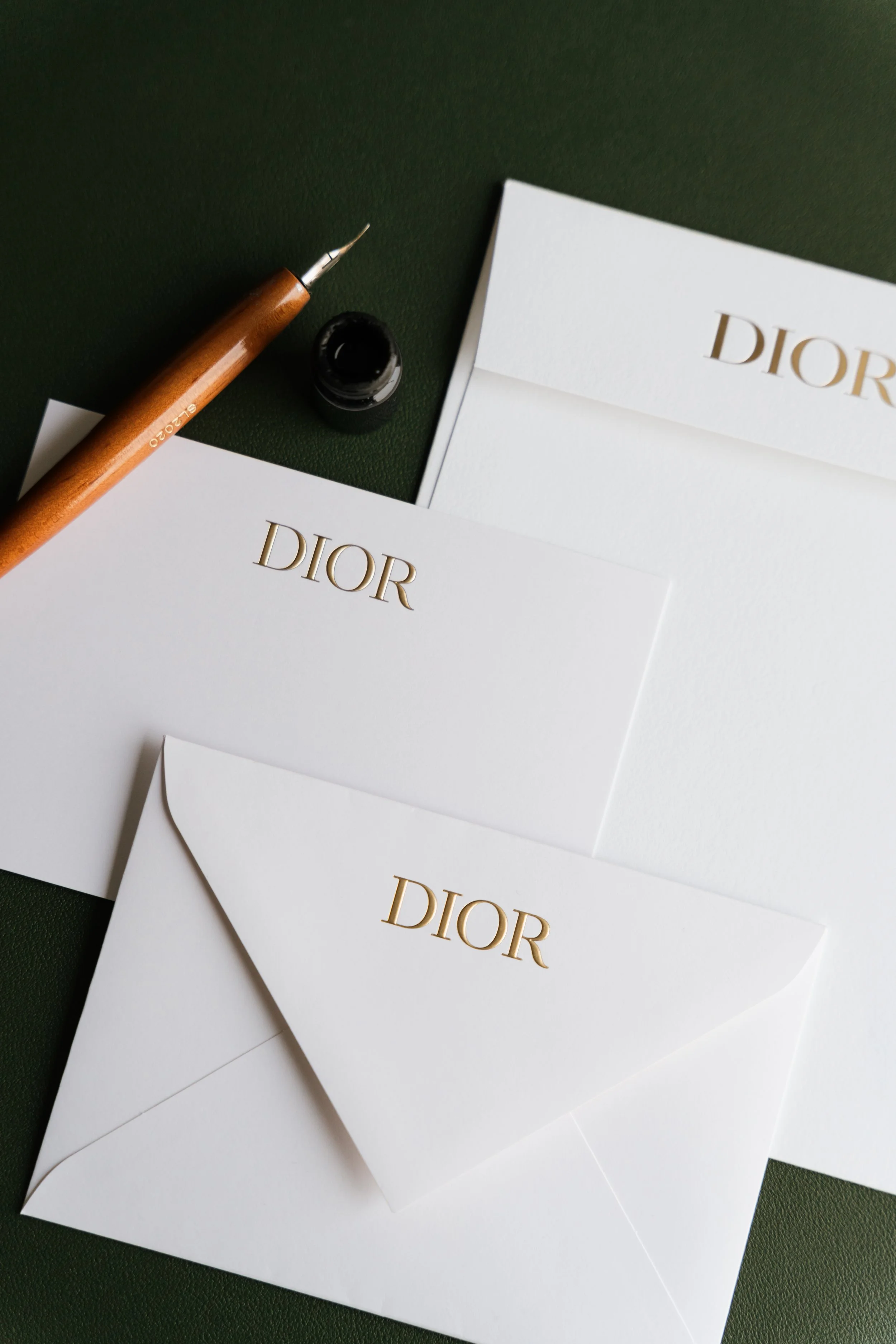 Multiple white envelopes with gold 'DIOR' logo, an ink pen with a wooden handle, and a black ink bottle on a dark green surface.