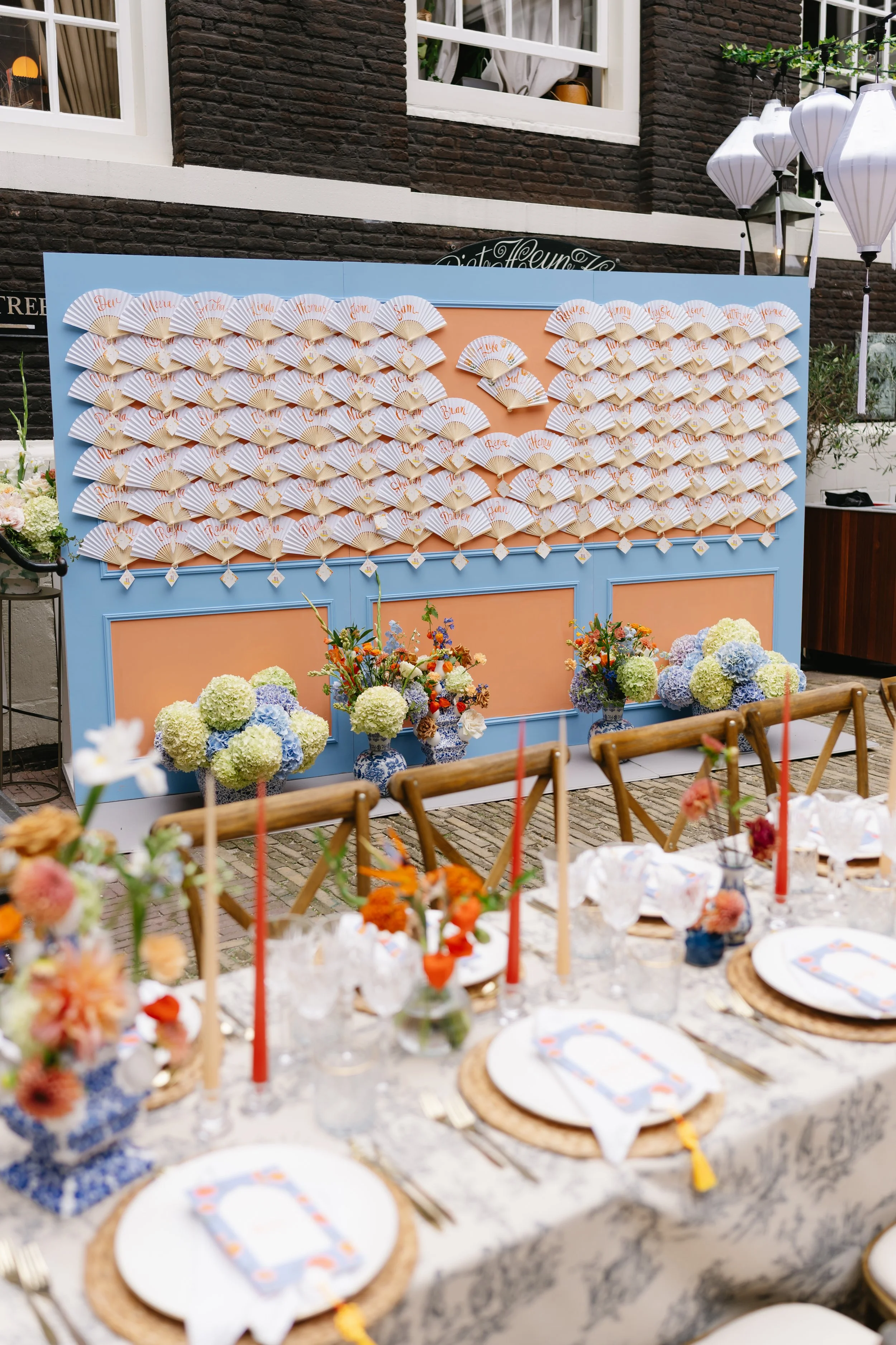 A decorated outdoor event space with a blue and peach backdrop featuring white fans with handwritten names, floral arrangements in blue and white vases, and a table set with plates, cutlery, and floral centerpieces.