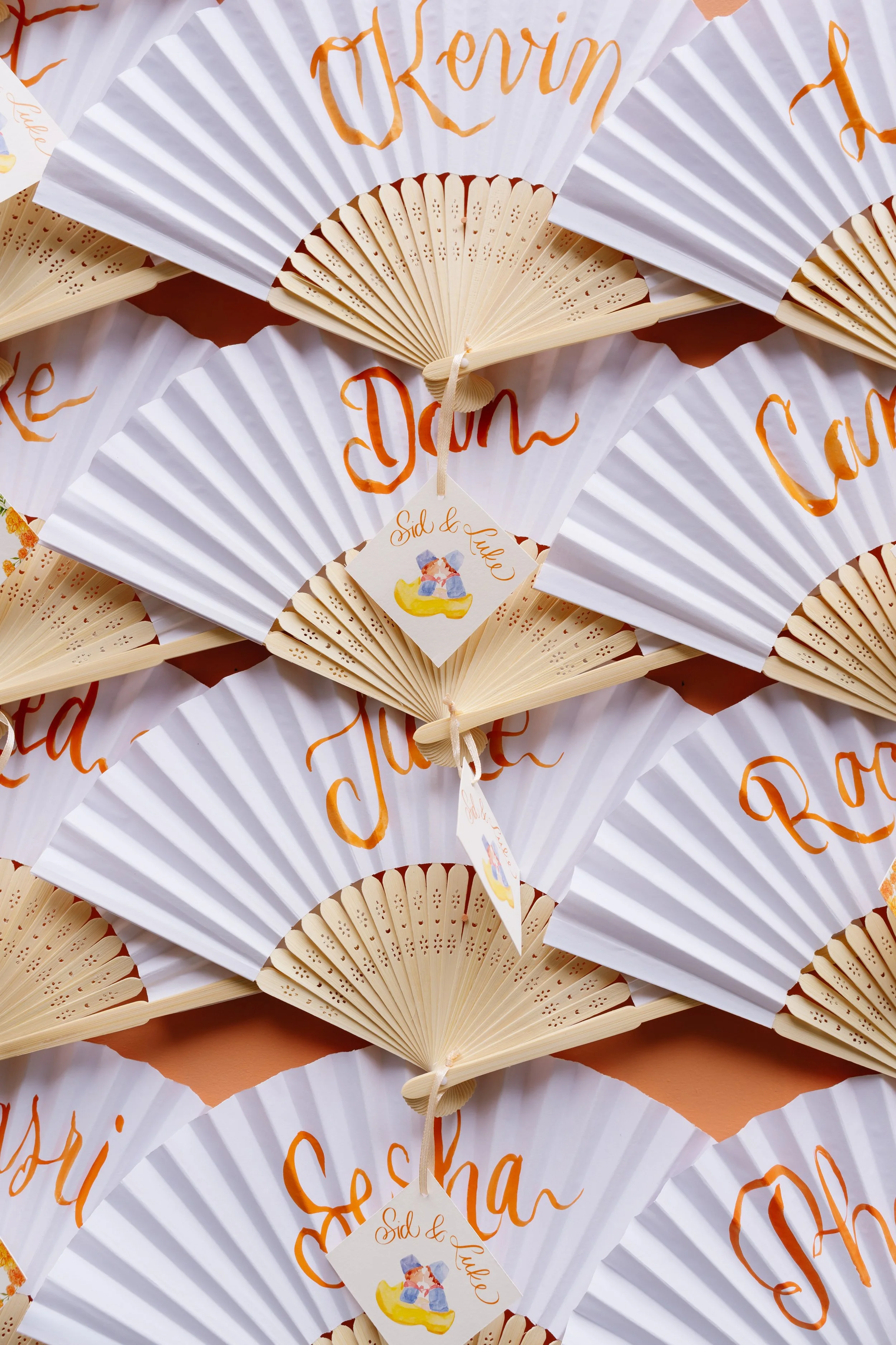 Multiple paper hand fans with white background, orange cursive writing, and a tag that reads 'Sid & Luke' with an illustration of two stuffed animals sitting on a yellow duck.