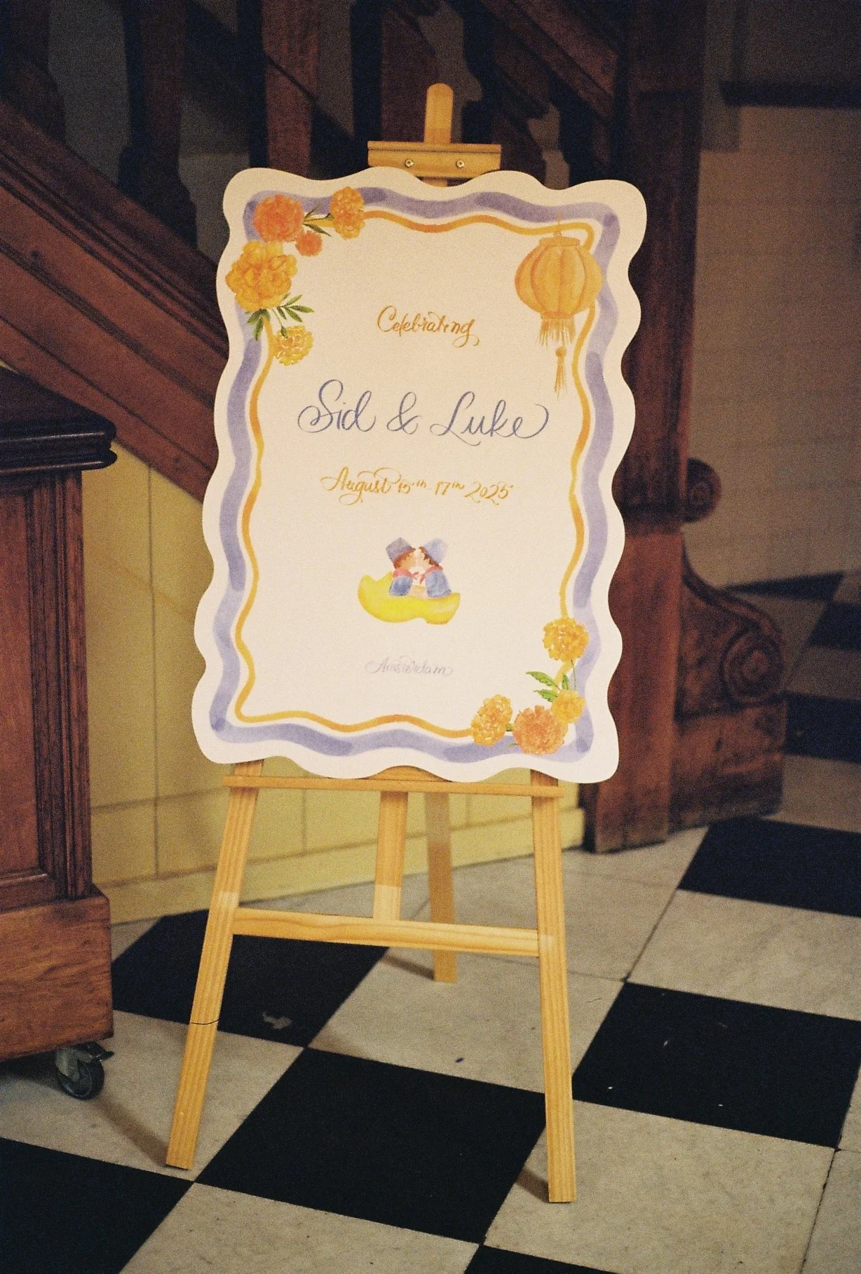 A decorative wedding sign on an easel with floral designs, a lantern illustration, and a couple illustration, announcing the celebration of Sid and Luke on August 16th, 2025, in Amsterdam.