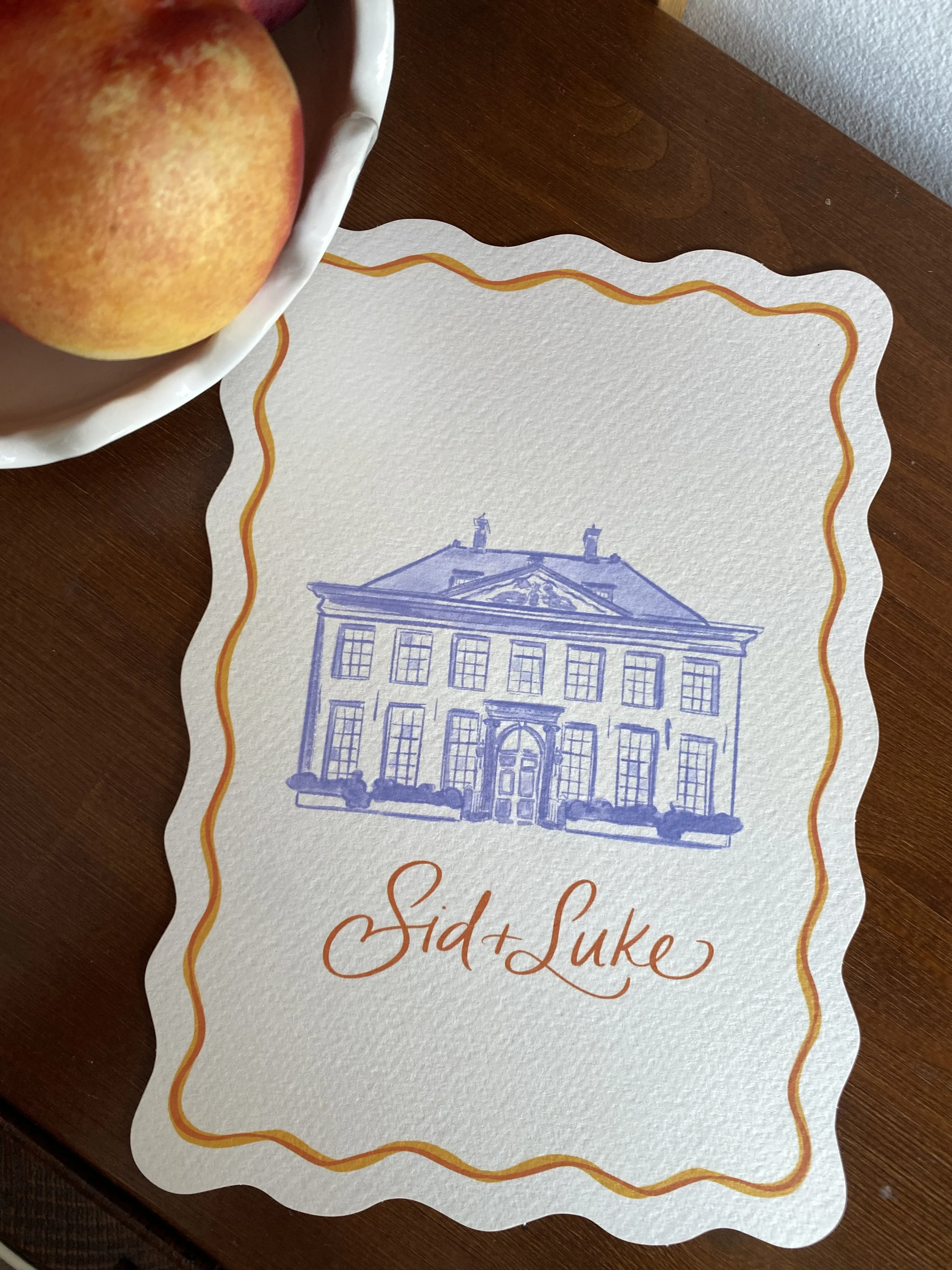 A paper with a decorative orange and yellow border, featuring a blue sketch of a building, and the text 'Sid & Luke' written in cursive red ink, lying on a wooden surface. Amsterdam Wedding
