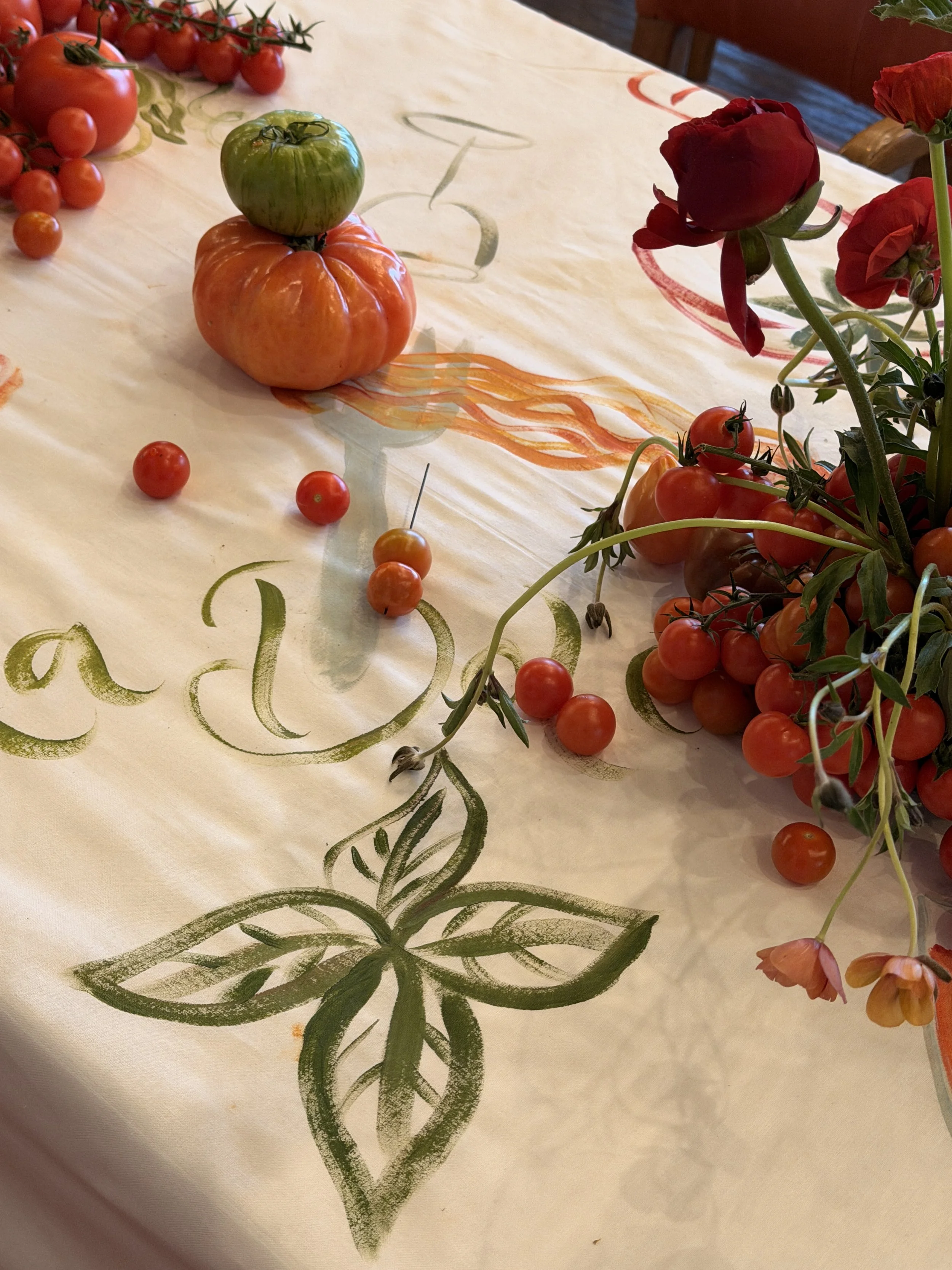 Decorative tablecloth with painted swirling green designs, topped with various tomatoes and red flowers.