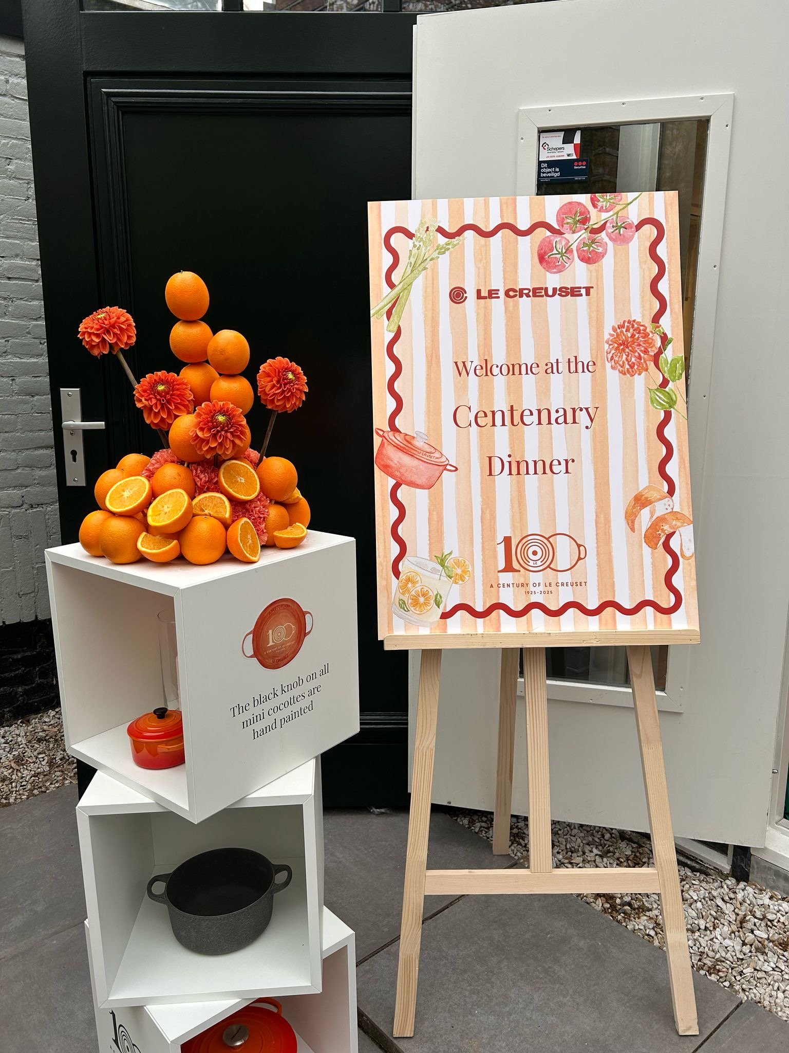 Decorative display of oranges and orange-themed flowers arranged in a pyramid, next to a sign celebrating 100 years of Le Creuset, with a white cabinet filled with kitchenware and a black pot on the bottom shelf.