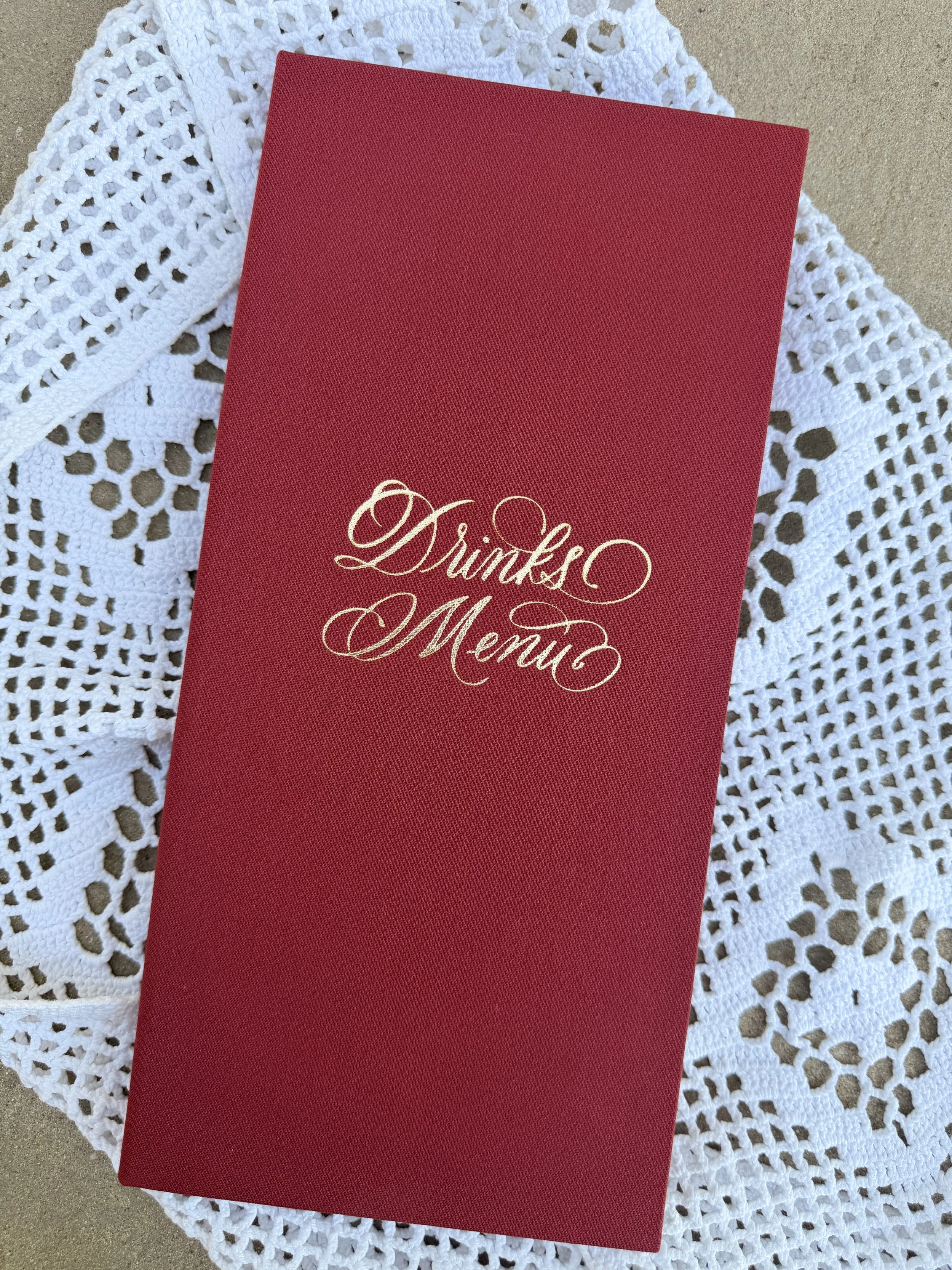 A red menu with gold embossed cursive text reading "Drinks Menu"  in a bespoke binder for wedding in Connemara - Ireland