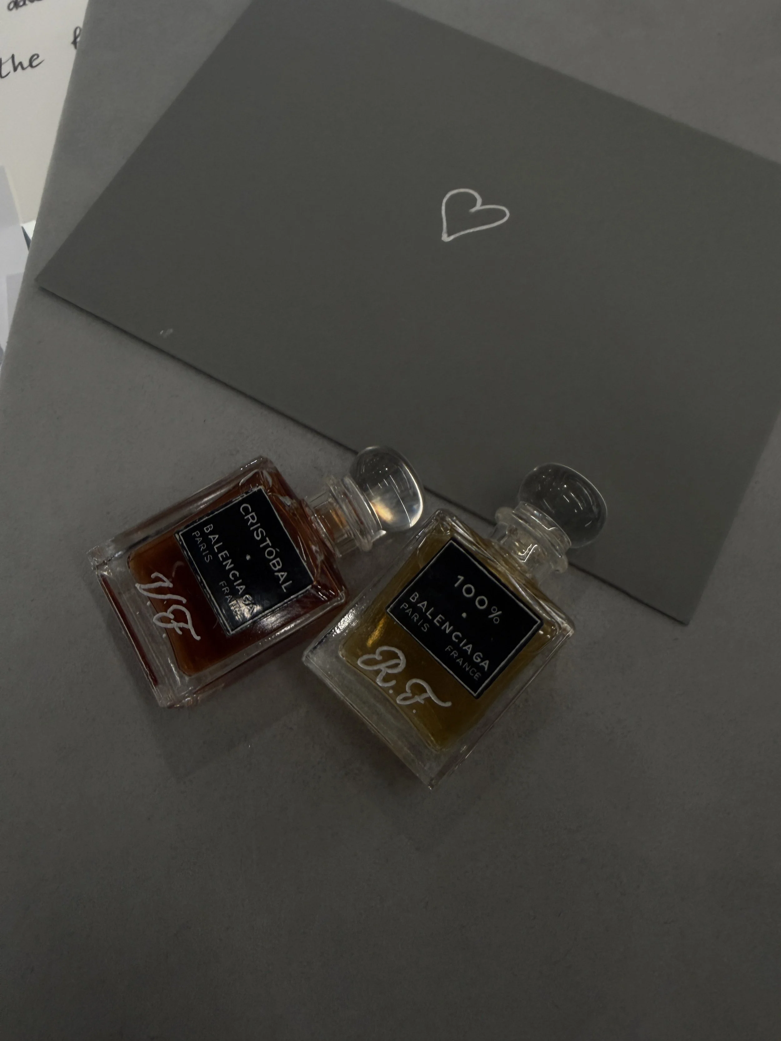 Two small perfume bottles labeled 'Cristobal Balenciaga Paris' and '100% Balenciaga Paris' on a gray surface with a gray box in the background featuring a white heart drawing.