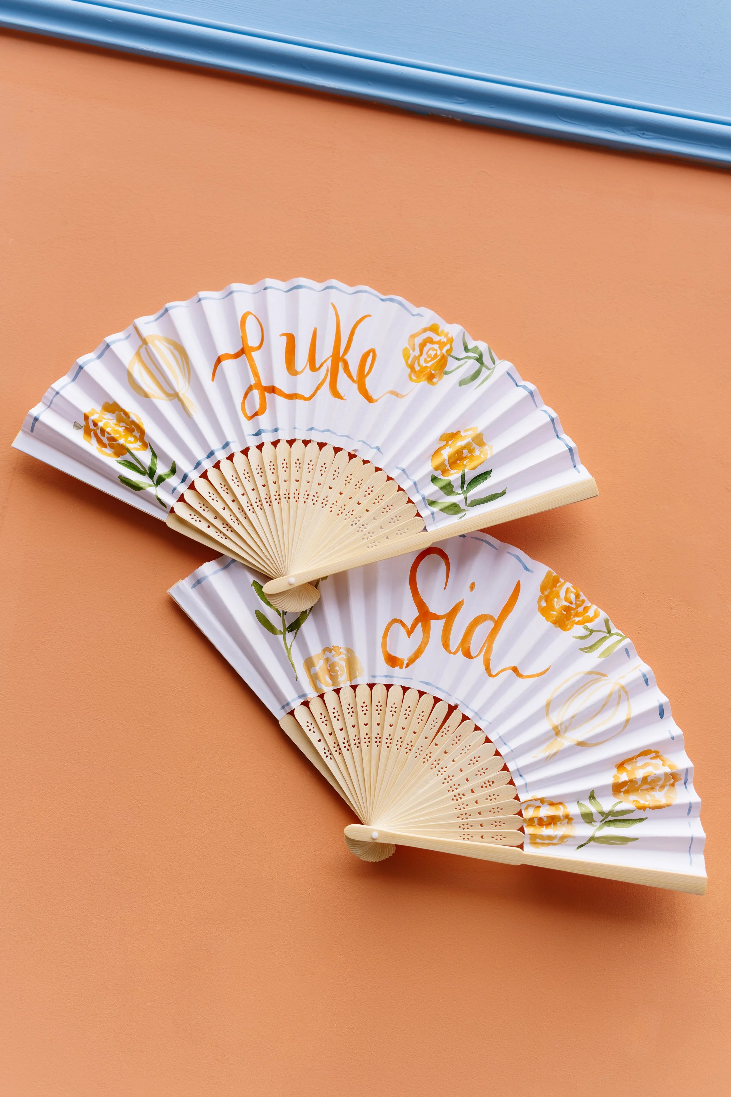 Decorative paper fan with the name calligraphy, floral designs, set against a peach-colored background with a blue trim at the top.