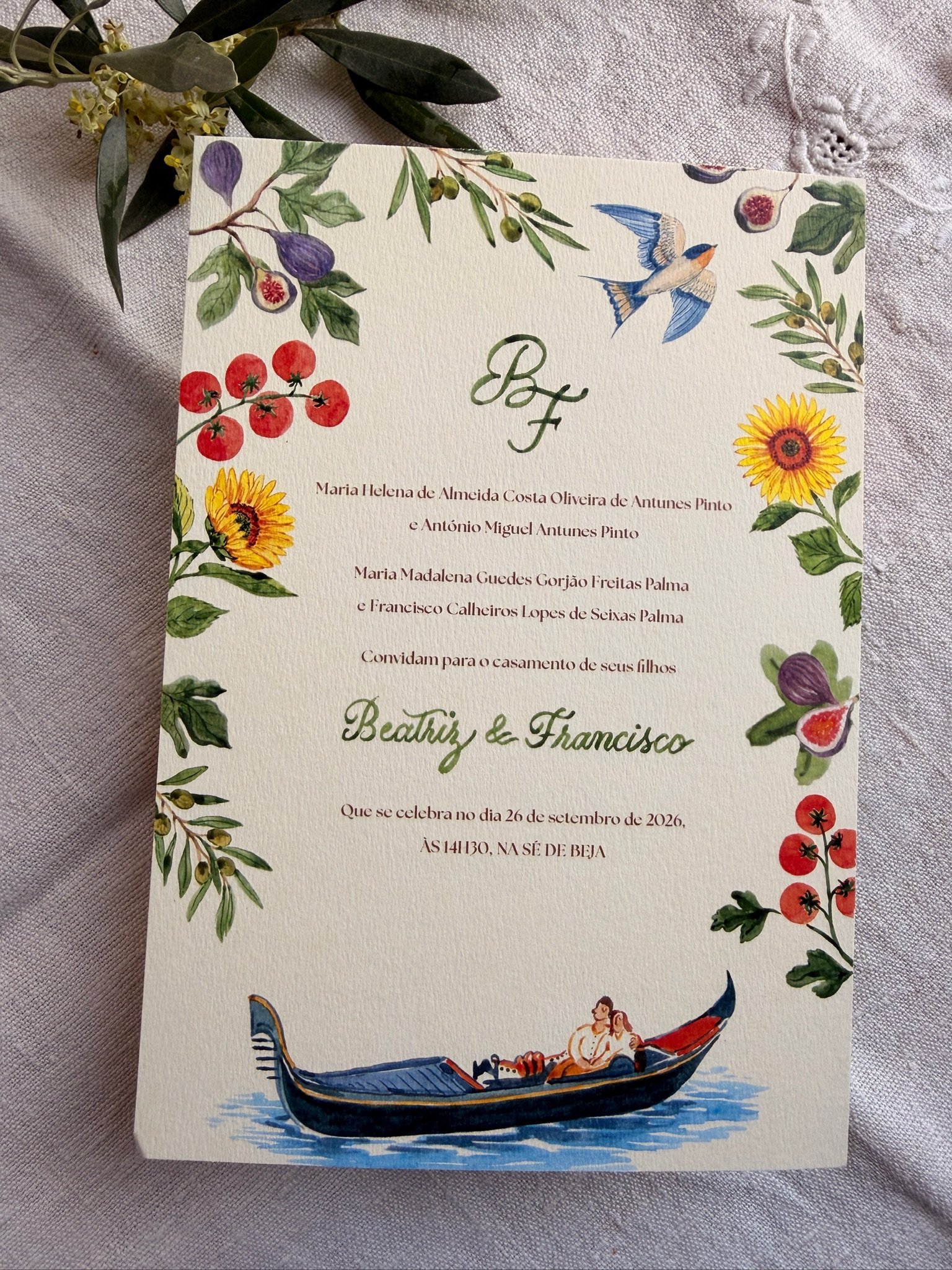 A wedding invitation with a floral border and watercolor illustrations of a boat, a swallow bird, figs, cherry tomatoes, and sunflowers on cream-colored textured paper. Portugal Wedding
