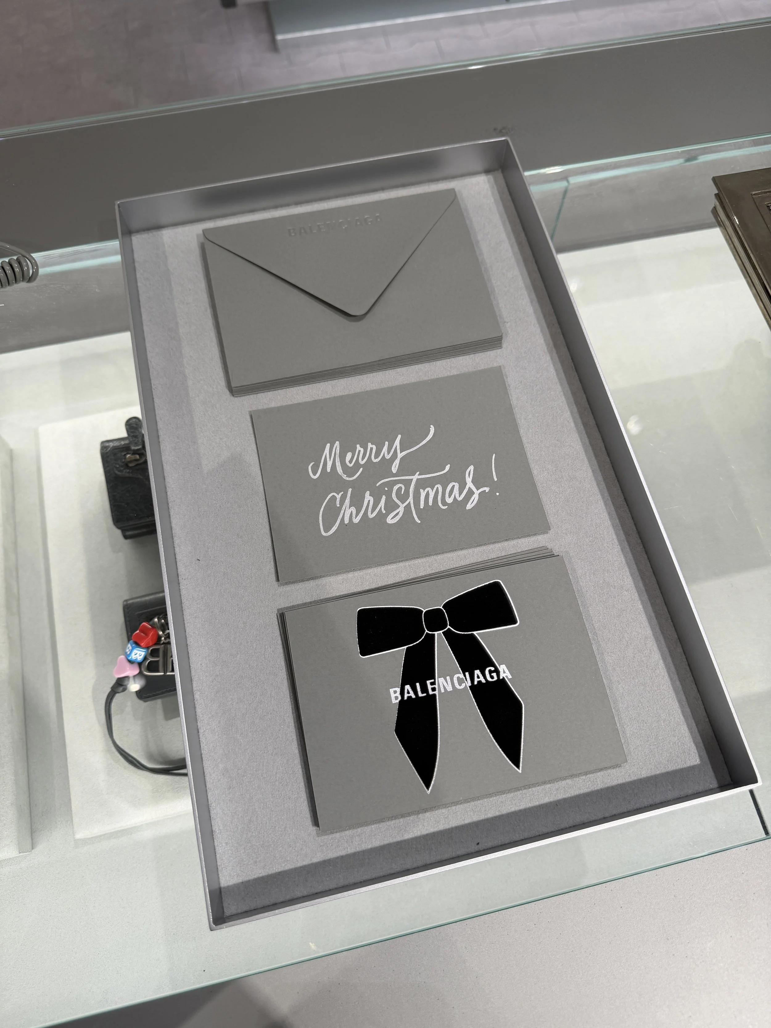 A display case containing grey greeting cards with festive messages and designer logos for Balenciaga, including a card with a black ribbon design.