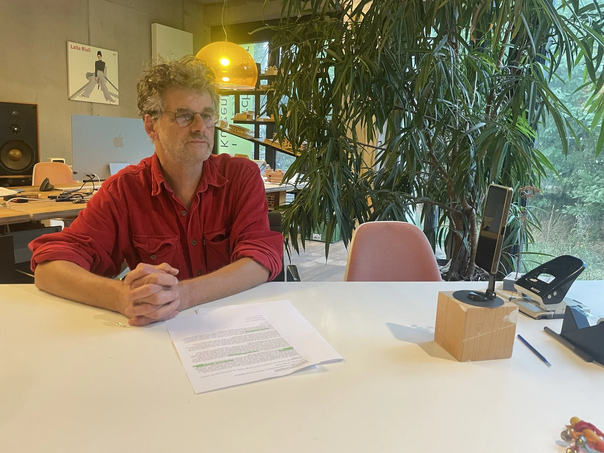 Ein Mann sitzt an einem weißen Tisch in einem modernen Büro. Vor ihm liegen Papiere und er trägt eine rote Jacke. Im Hintergrund sind eine große grüne Pflanze, ein Monitor, ein Lautsprecher und moderne Lampen. Es gibt auch eine Fensterfront, die viel Tageslicht hereinlässt.