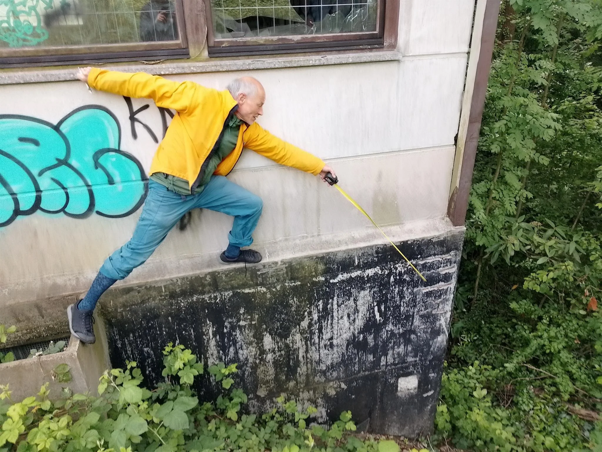 Ein älterer Mann in gelber Jacke, der auf einer Mauer steht und sich nach unten beugt, während er eine Taschenlampe hält. Die Wand ist mit Graffiti bemalt und es gibt Grünpflanzen im Vordergrund.