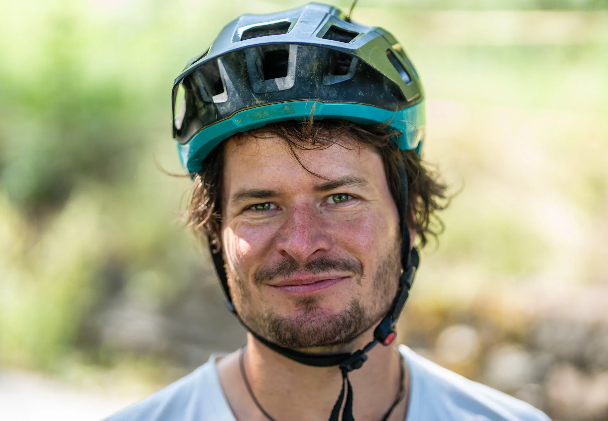 Ein Mann trägt einen schwarzen Fahrradhelm mit blauen Akzenten und hat braune, lockige Haare, lächelt und steht im Freien mit grünem, unscharfen Hintergrund.