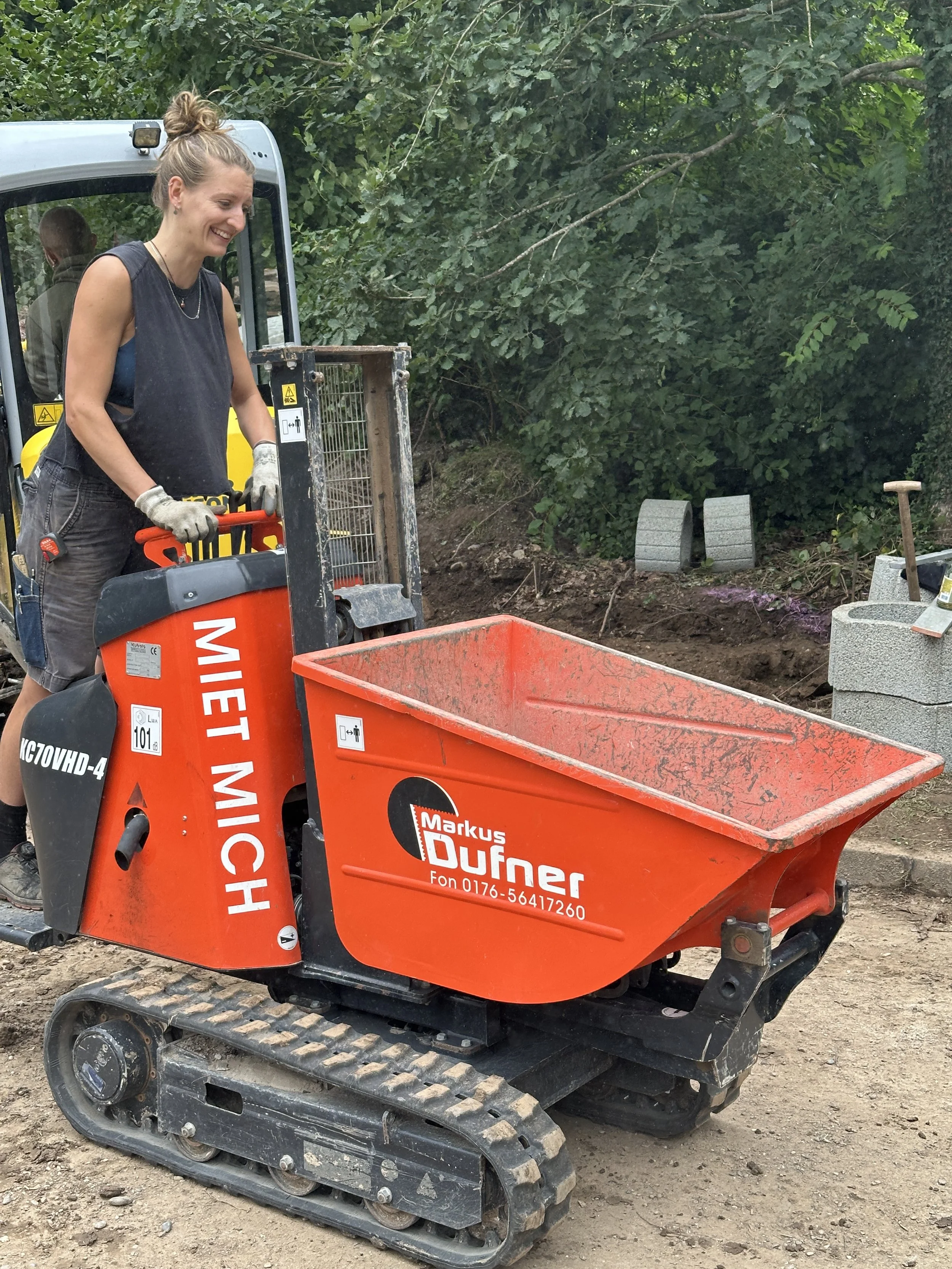 Ein lachender Mensch steuert einen kleinen Spielzeug-Bagger auf einer Baustelle, mit Büschen im Hintergrund und Baumaterialien am Boden.