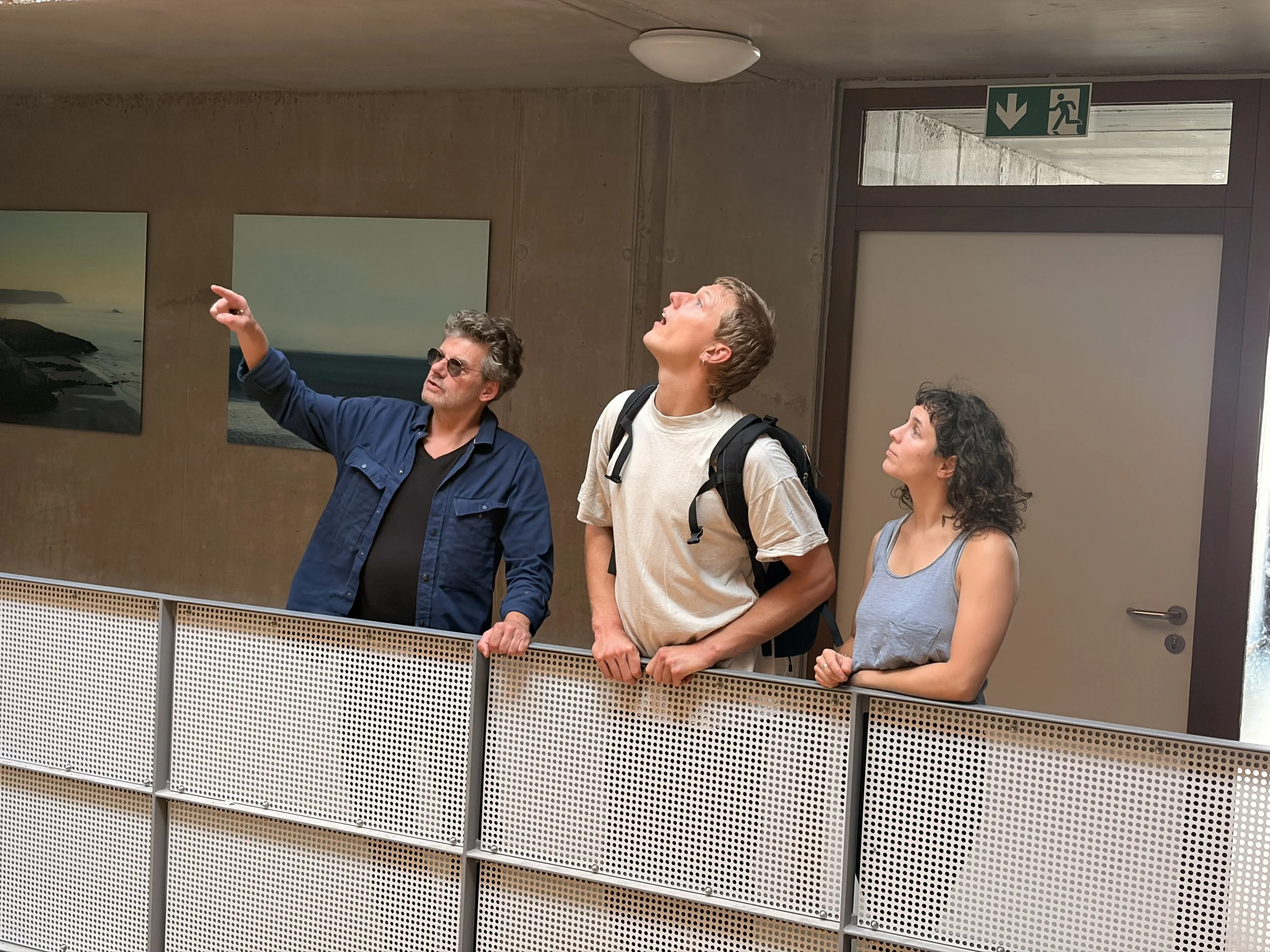 Drei Menschen stehen an einer Geländer an einer Wand, beobachten etwas und diskutieren. Der Mann auf der linken Seite zeigt nach oben, während die anderen beiden nach oben schauen.