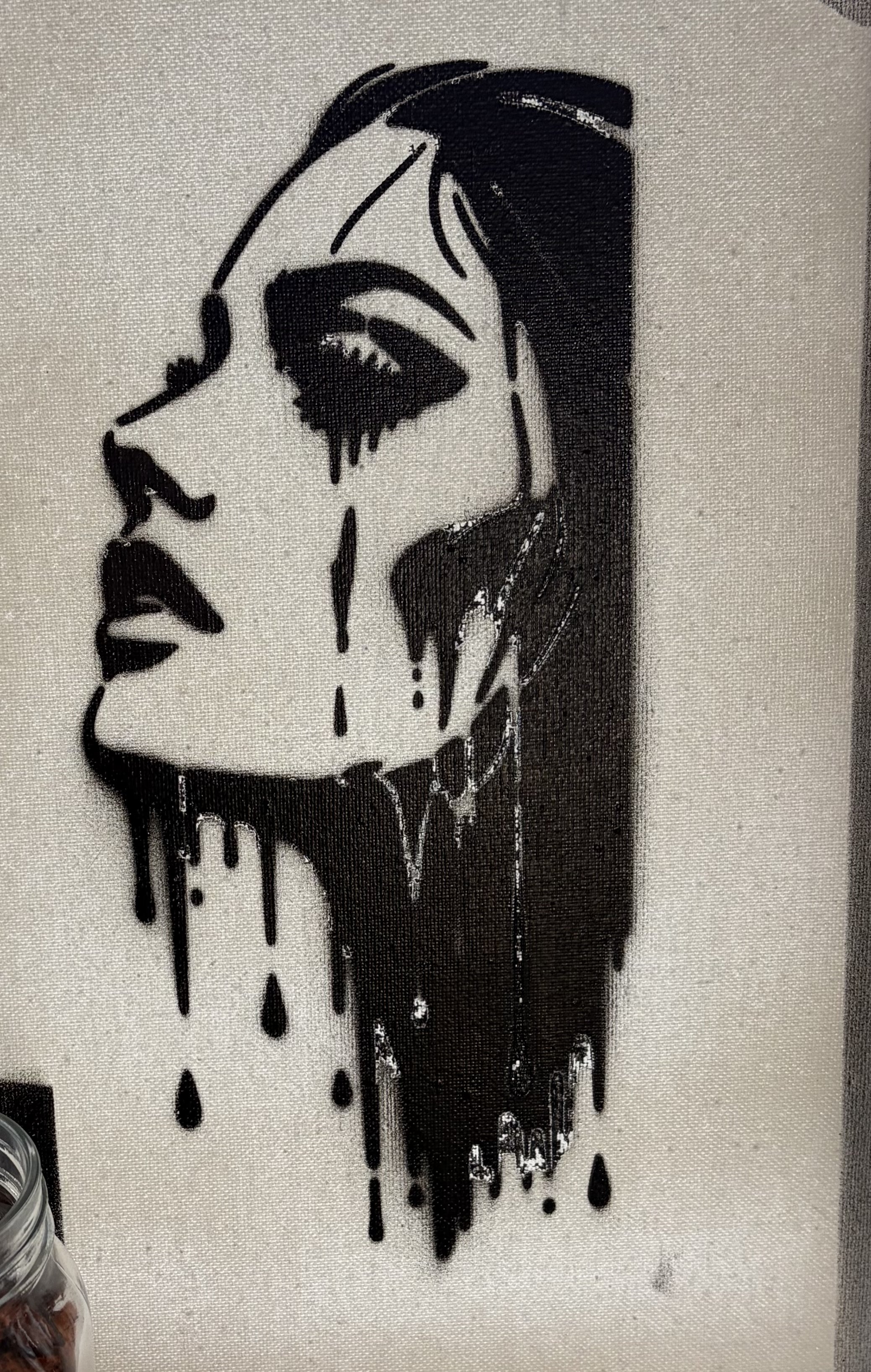 Black spray paint stencil of a woman's profile with makeup, dripping paint effect, on a white textured surface.