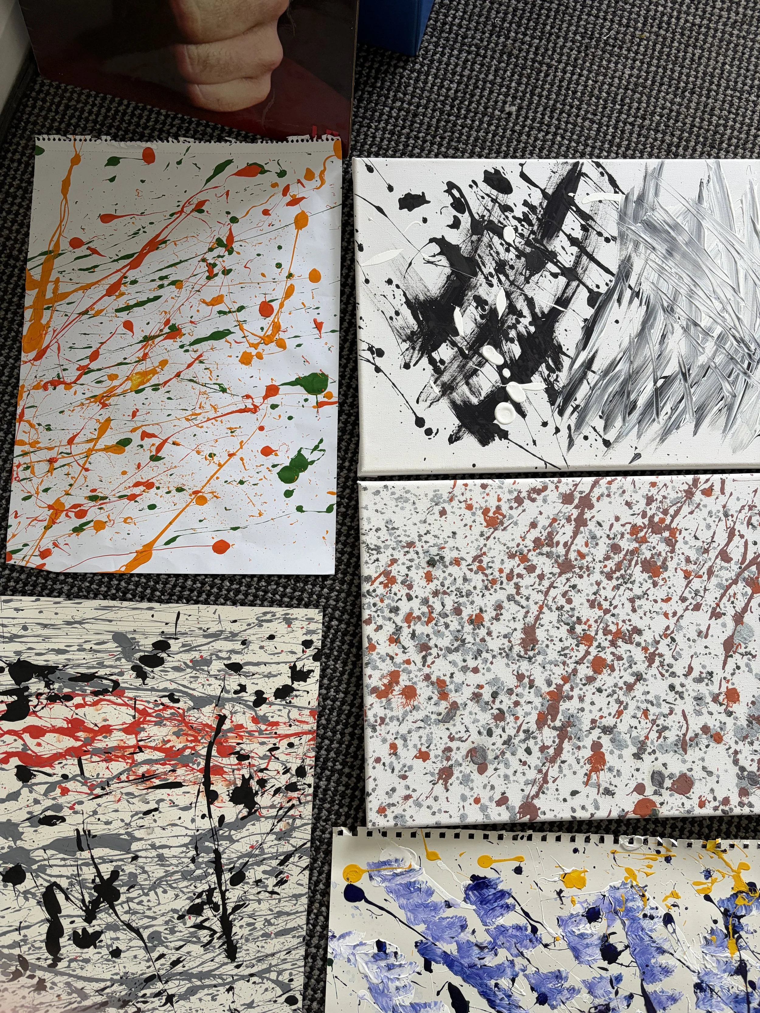Assorted abstract paintings on canvases with splatters and streaks of paint in various colors, including black, red, orange, green, blue, and gray, displayed on a carpeted floor.