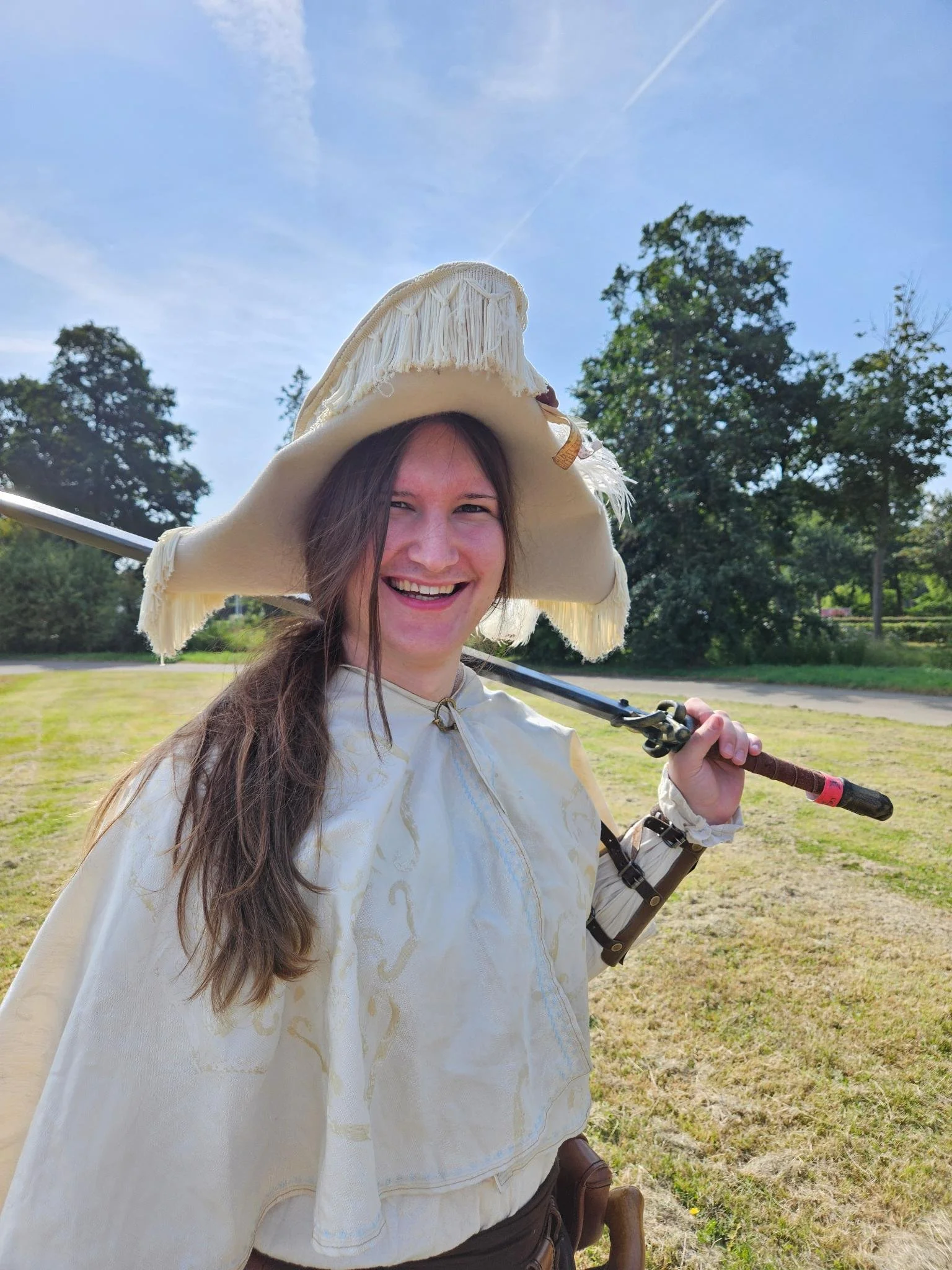 A photo of a white brunette, her hair in a messy ponytail. She is standing in a field, trees behind her. The woman is wearing an off-white halfcloak embroidered with gold, a white tunic, vambraces, a cream admiral's tricorn, and is holding a sword.